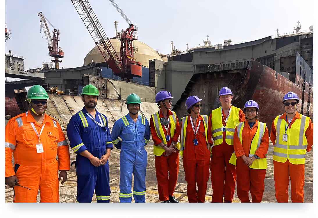Group of workers in helmets and high-visibility gear standing in front of shipyard with large ship under construction.
