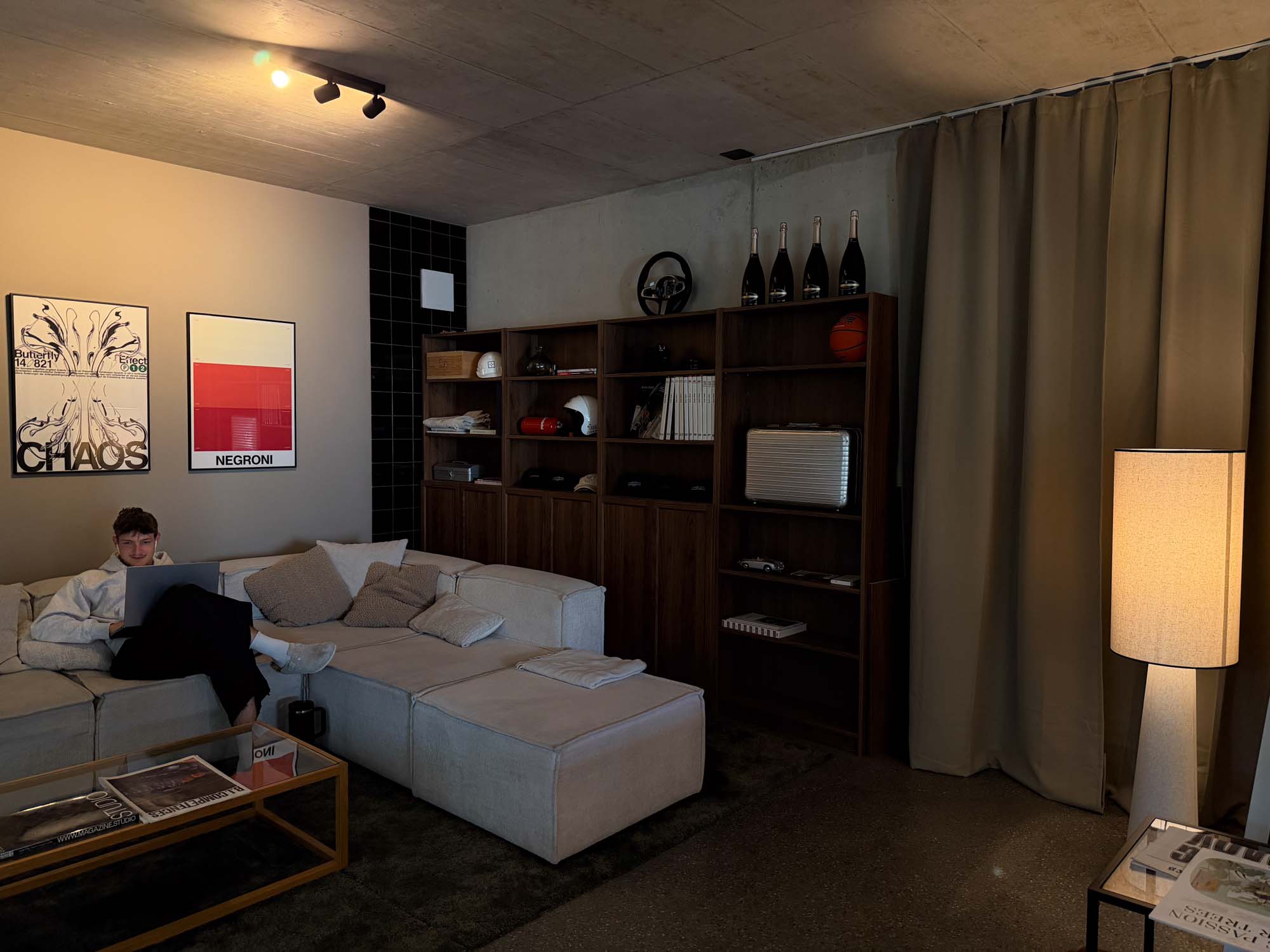 A modern living room featuring a neutral-toned sectional sofa, wooden bookshelf with decorative items, and a standing lamp. A person is sitting on the sofa, using a device. The atmosphere suggests a creative workspace, suitable for a Performance Marketing Agency in Zurich.