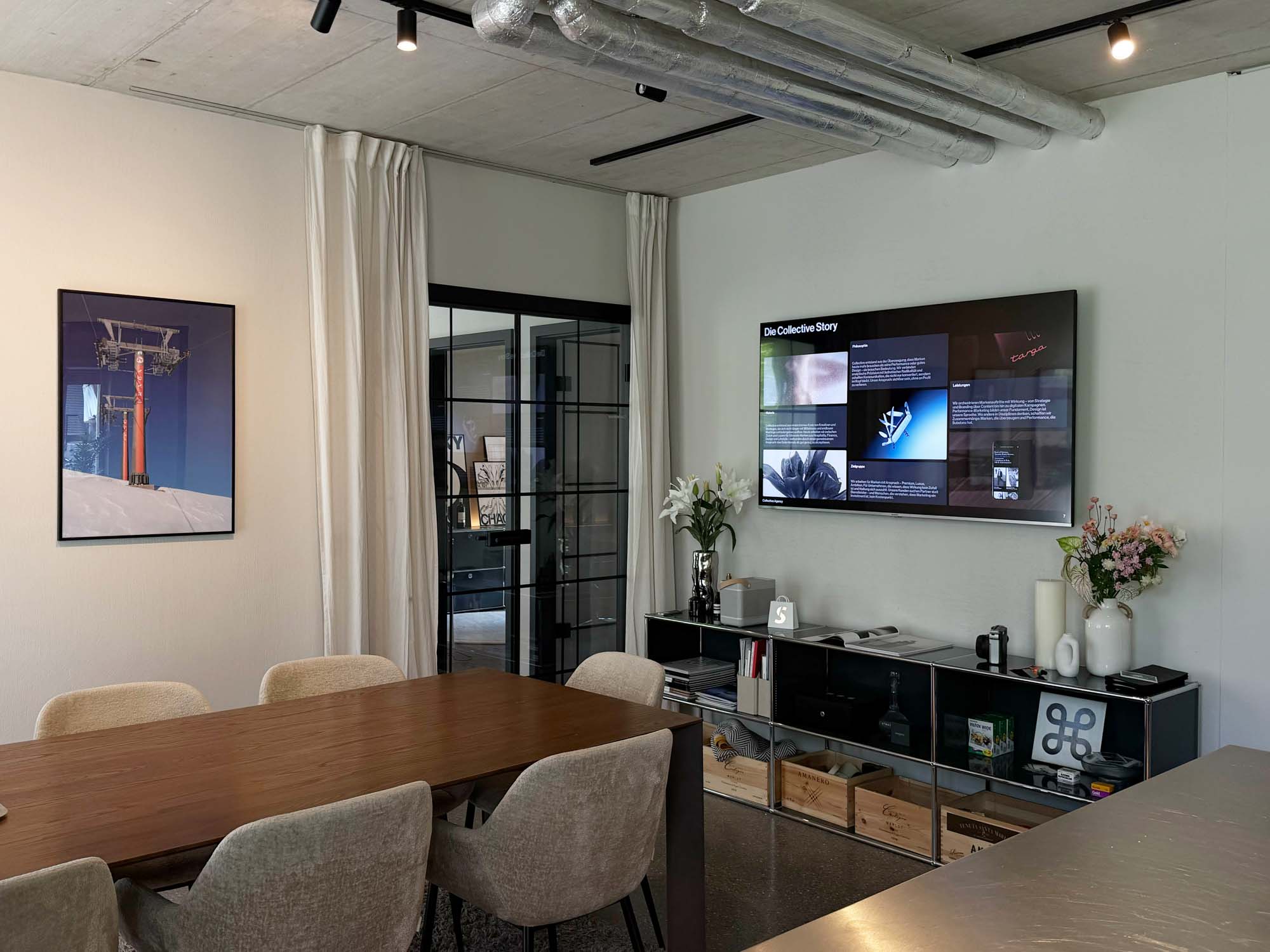 A modern office space featuring a wooden dining table with light-colored chairs, a large TV displaying digital content, a potted plant, and a framed image of a ski lift on the wall. This setting reflects a professional environment for a performance marketing agency located in Zurich.