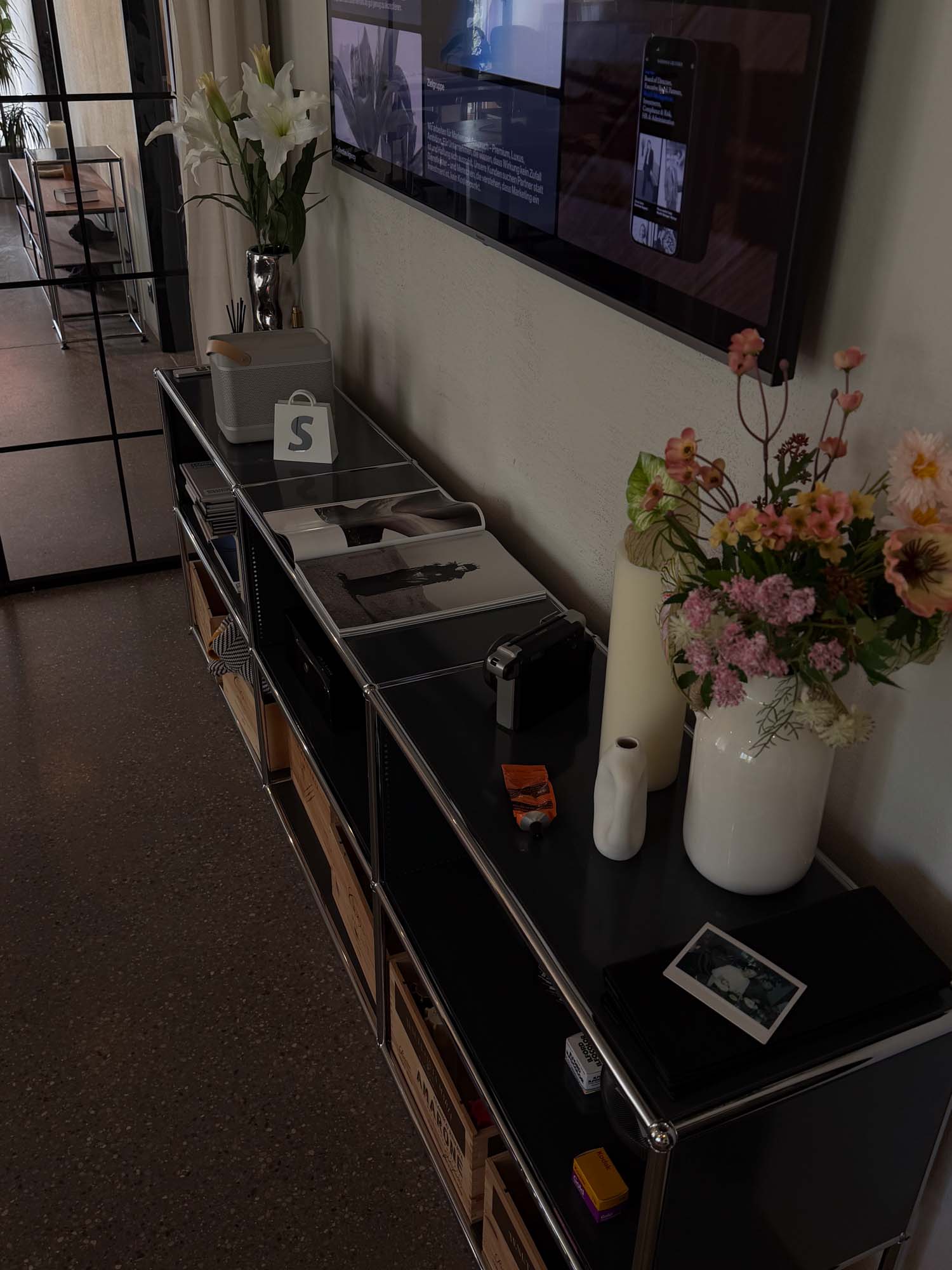 An interior shot of a modern office with a side table displaying flowers, a vase, a candle, and framed photos. A digital screen is mounted on the wall, showing various visuals, typical of a Performance Marketing Agency in Zurich.
