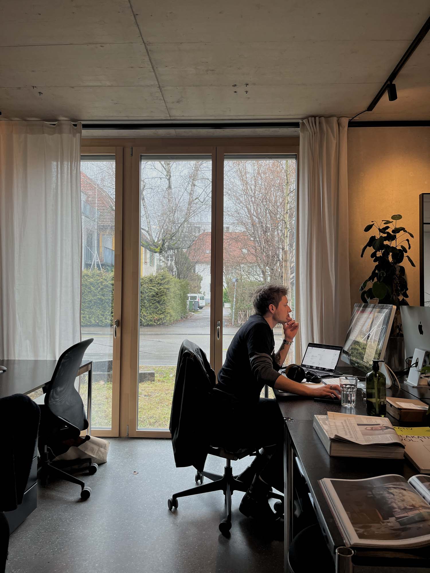 A man working at a desk in a modern office setting, seated in front of a computer. Natural light flows through large windows, revealing greenery outside. The workspace is organized, with a plant and some documents visible. This scene relates to a Performance Marketing Agency in Zurich.