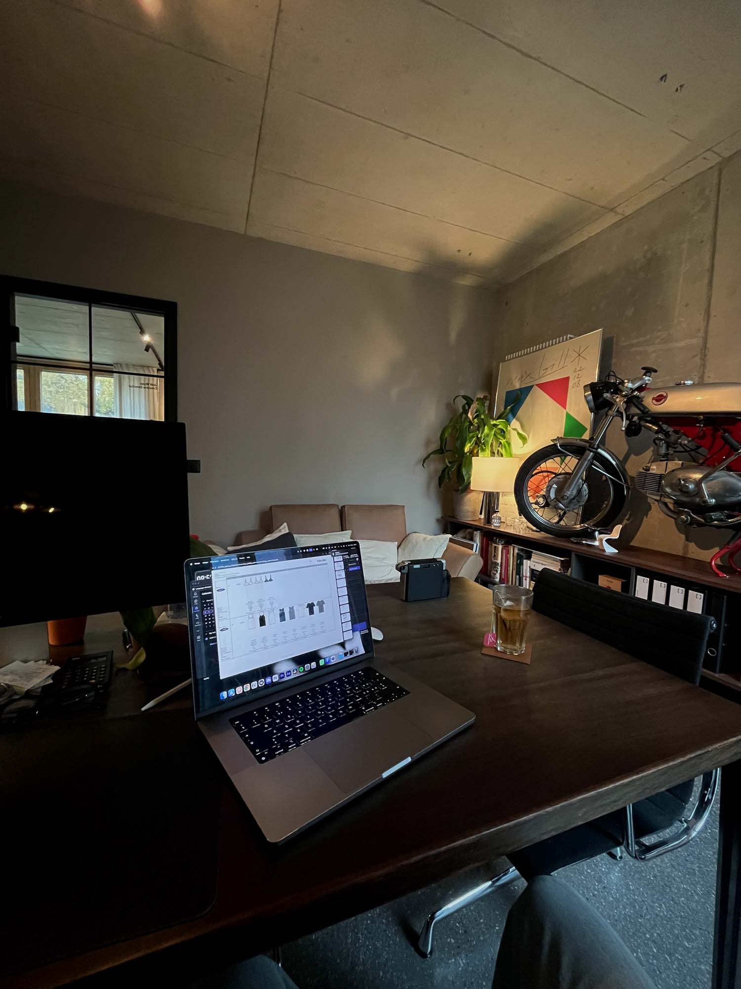 A workspace featuring a laptop open to a digital interface, a large monitor, and a drink on a dark wooden table. In the background, there is a couch, a plant, and a motorcycle mounted on a wall, suggesting a creative Performance Marketing Agency setting in Zurich.