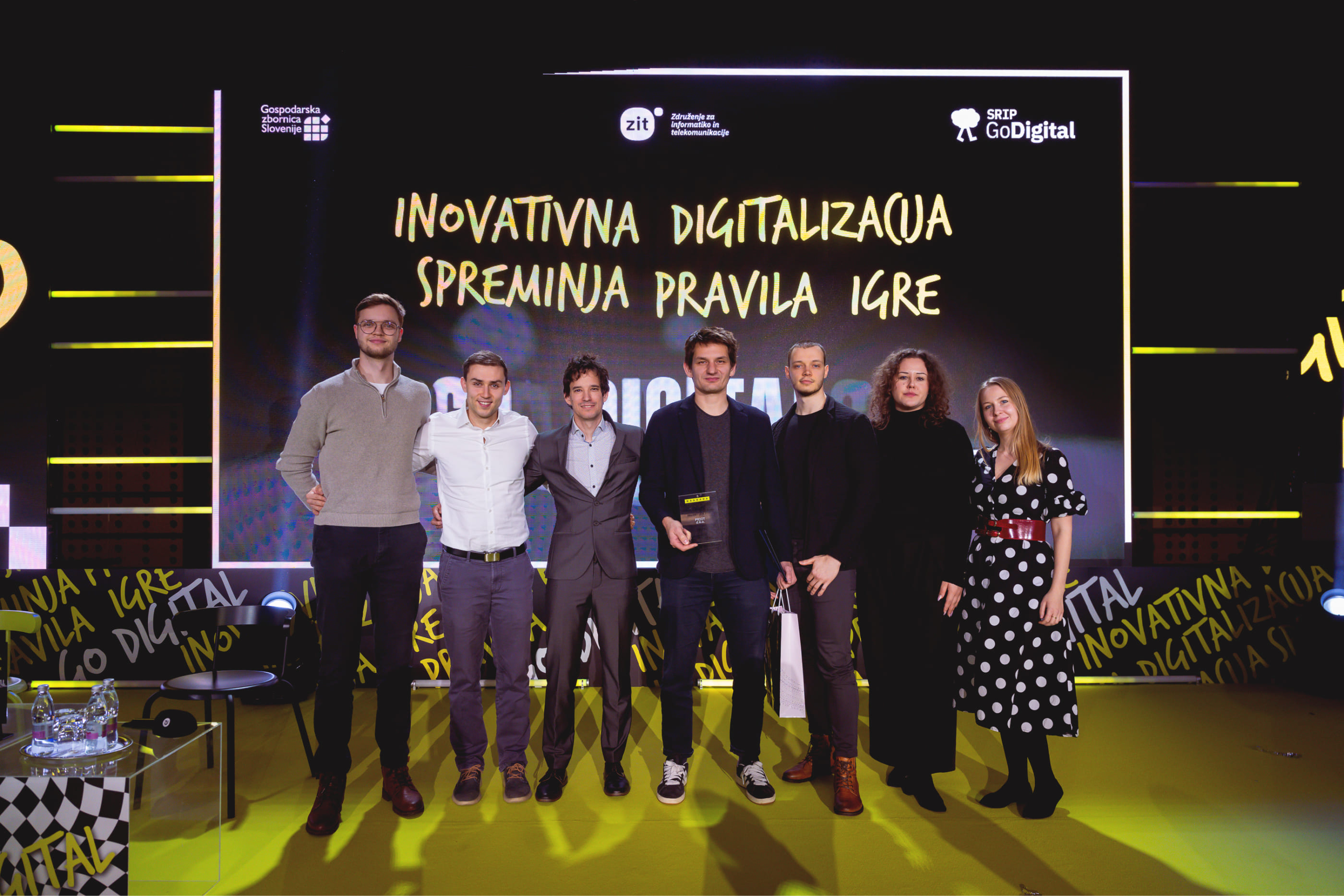 Group of seven people standing on a yellow stage in front of a screen with Slovenian text about innovative digitalization.