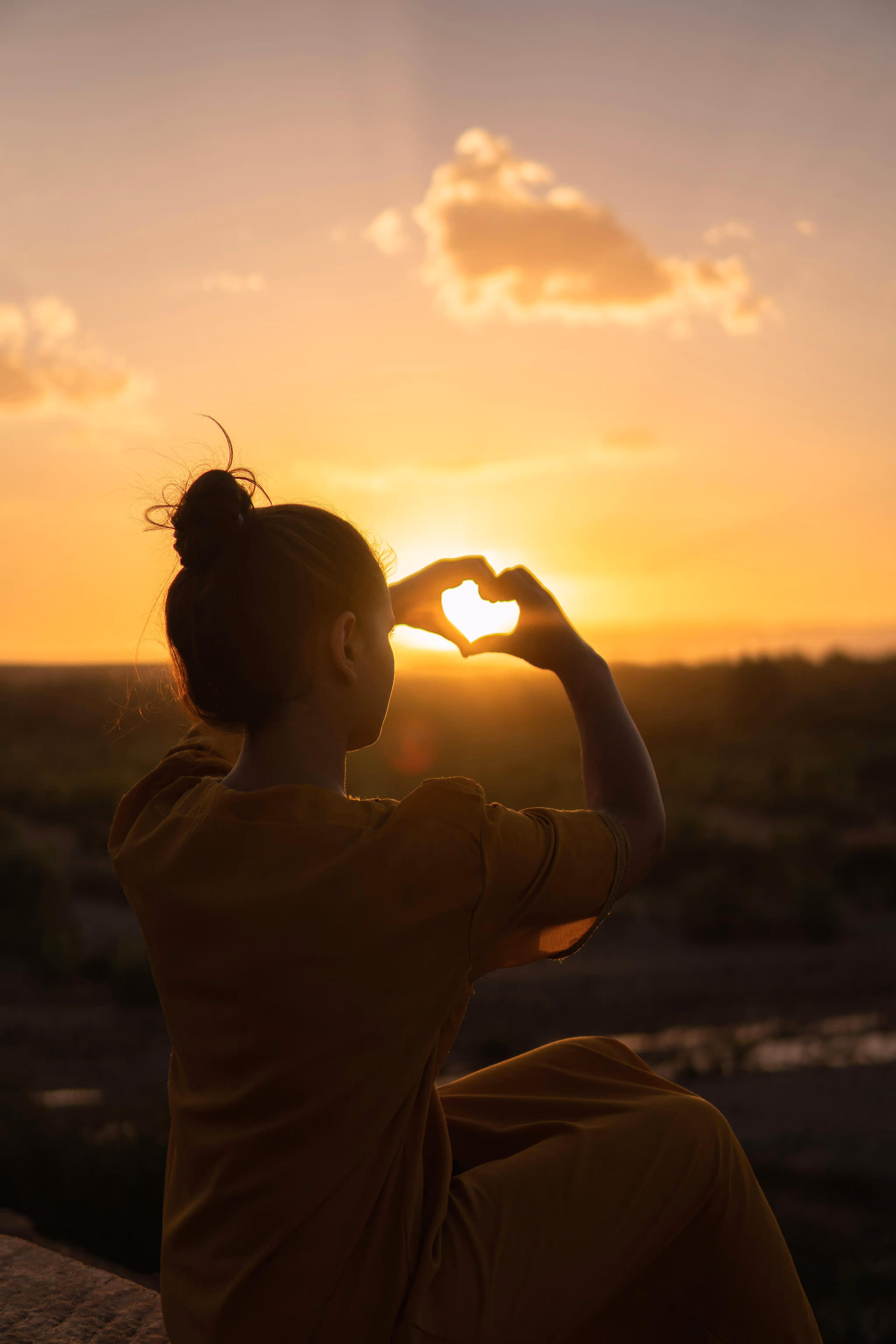 A person sitting on a ledge at sunset, forming a heart shape with their hands, silhouetted against the colorful sky.