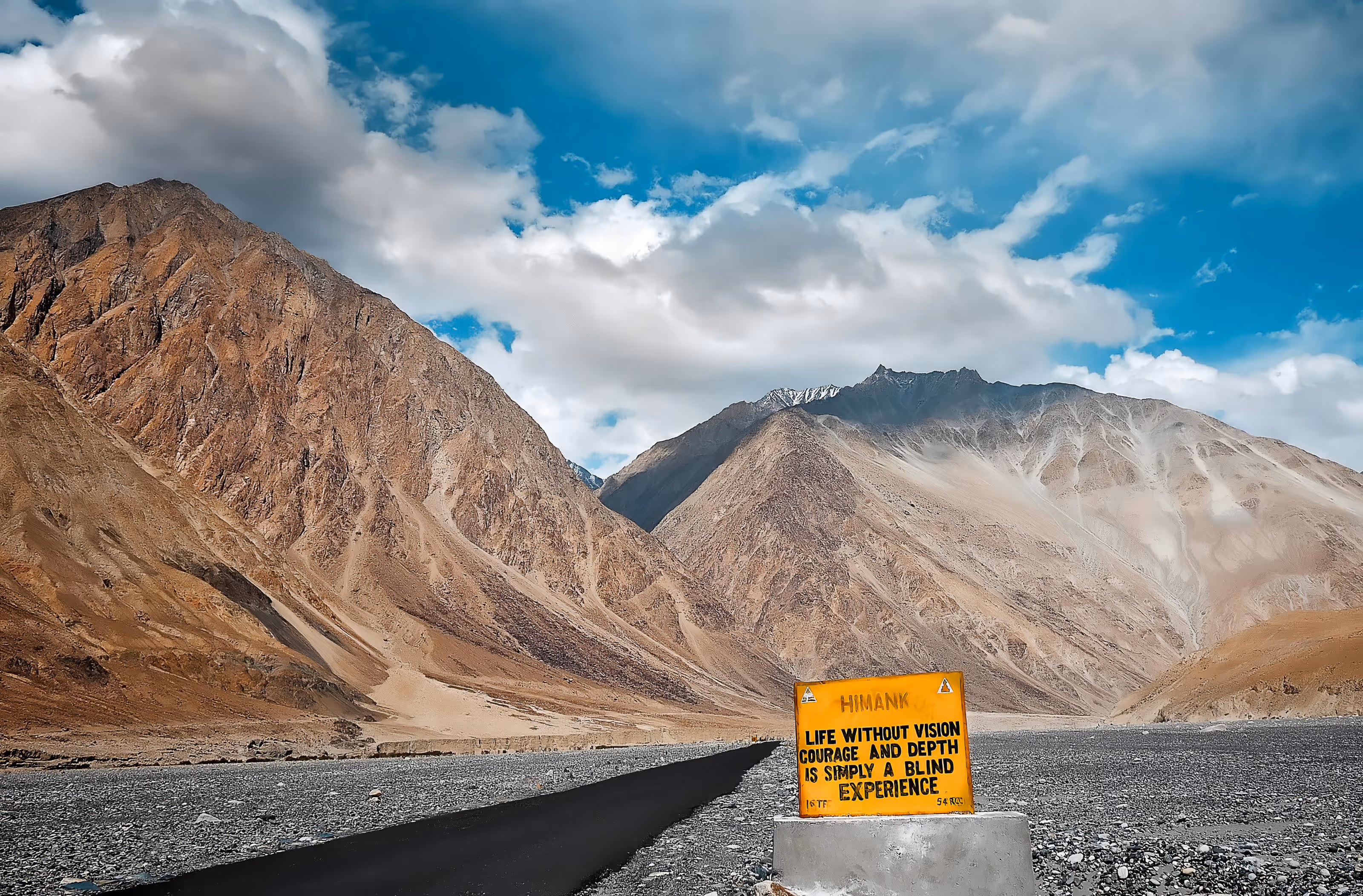 A winding road leads through a mountainous landscape, with rocky slopes and a blue sky filled with clouds. A warning sign on the side of the road states, 'Life without vision, surface and depth is simply a blind experience.'