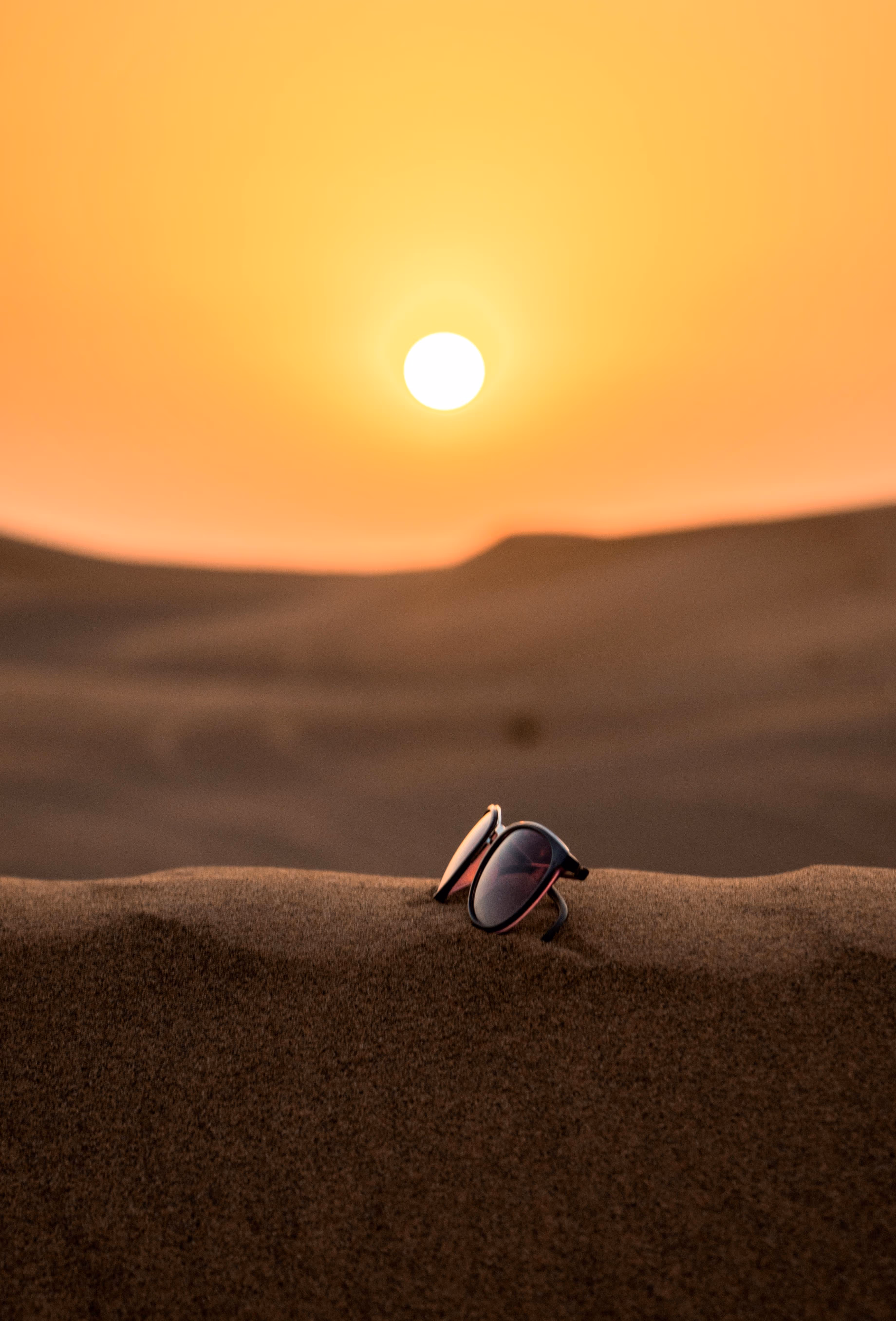 Sunglasses resting on sand dunes with a large sun setting in the background, illuminating the sky with warm orange hues.