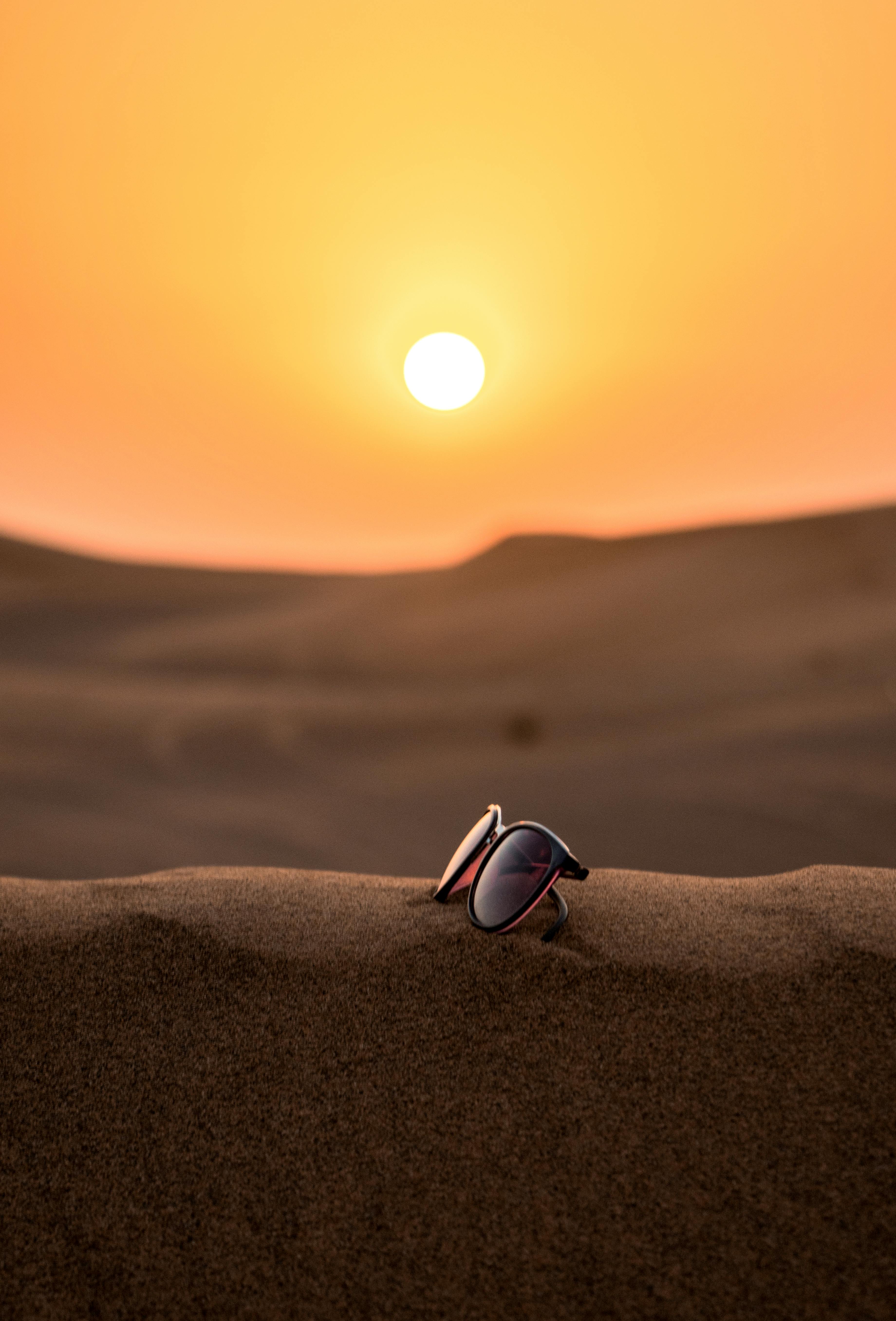 Sunglasses resting on sand dunes with a large sun setting in the background, illuminating the sky with warm orange hues.