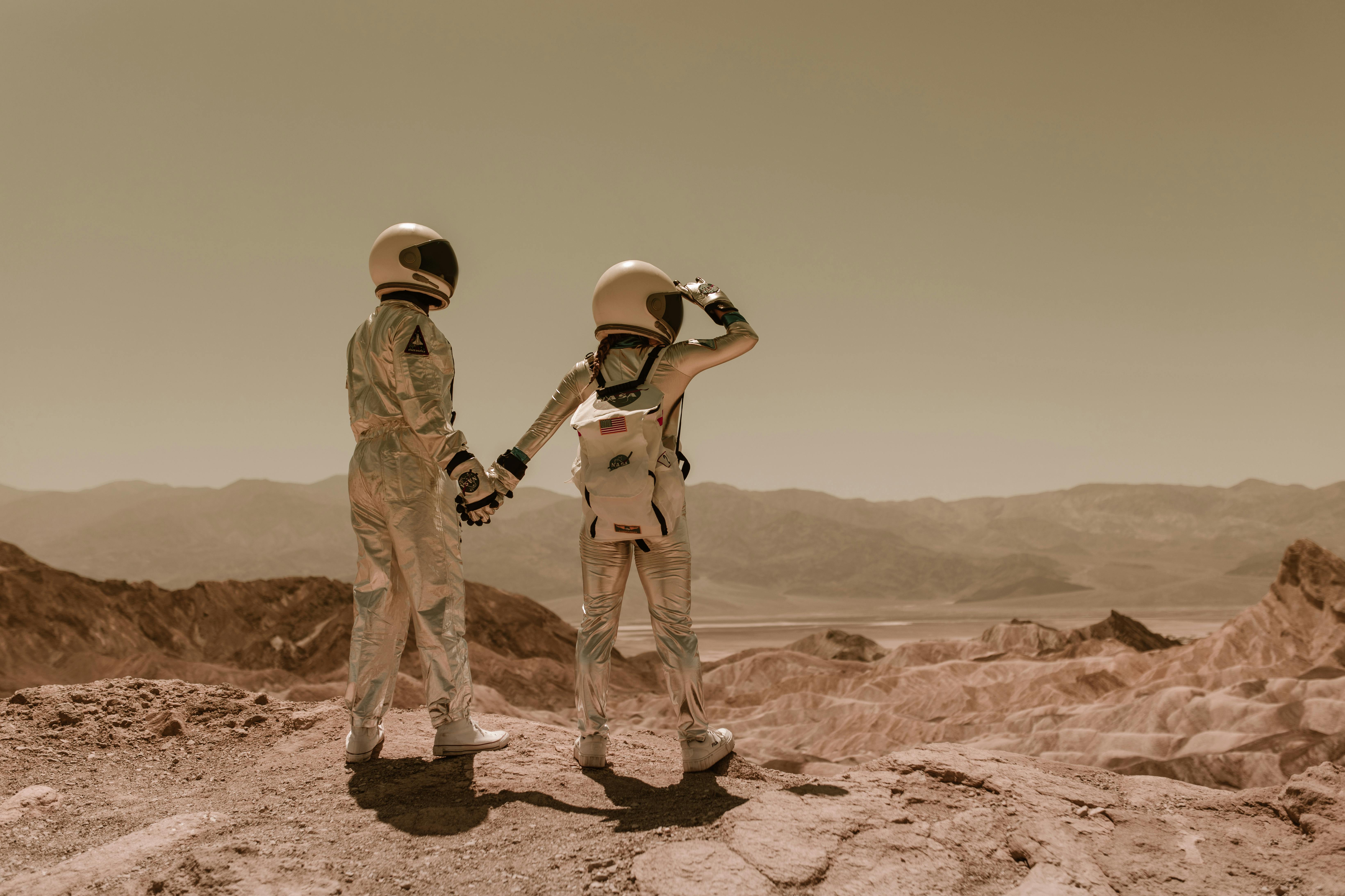 Two astronauts holding hands while standing on a rocky surface, looking at distant mountains under a hazy sky. The scene evokes a sense of exploration and adventure.