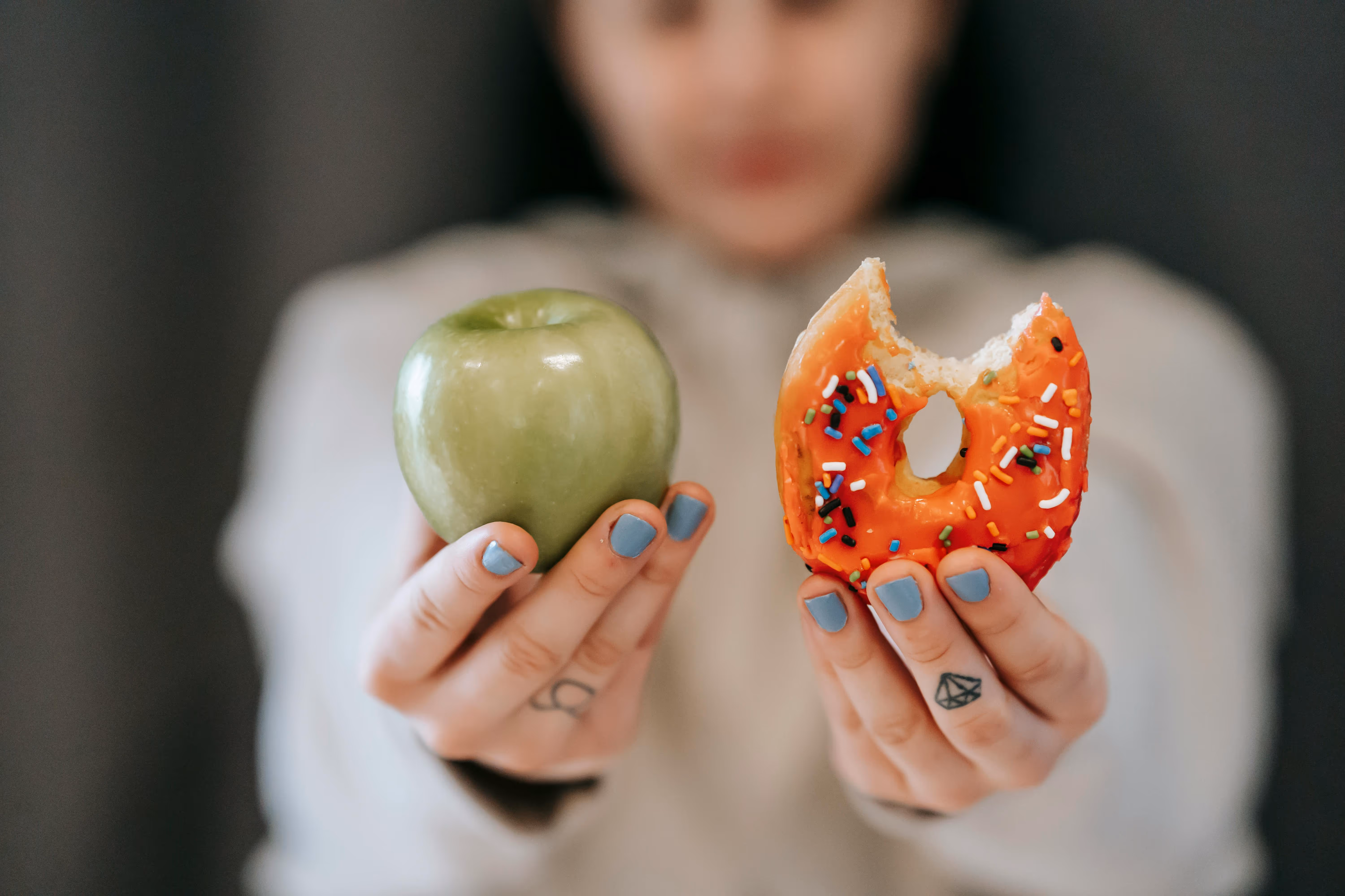 A person holding a green apple in one hand and a partially eaten donut with colorful sprinkles in the other hand, with a blurred background.