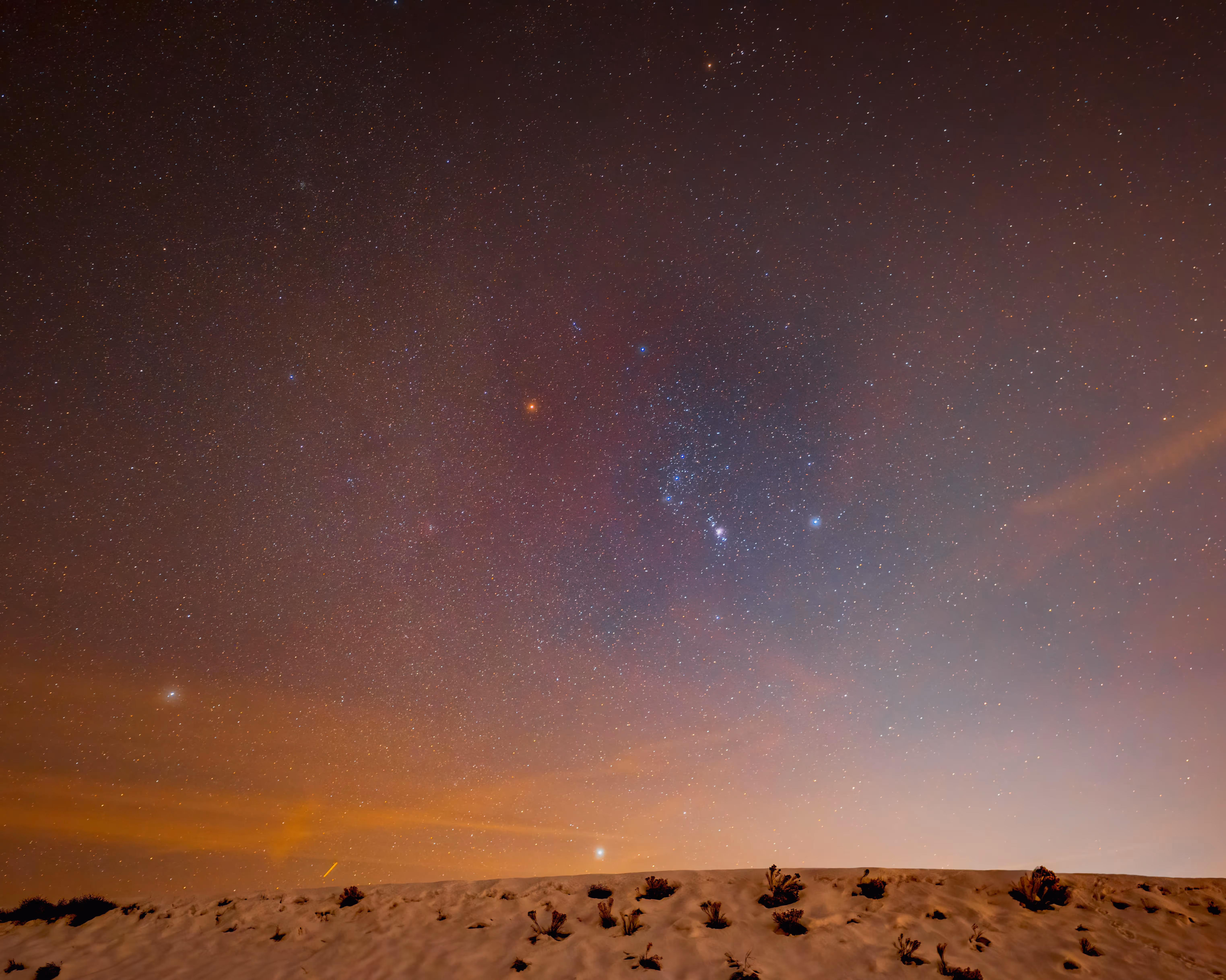 A night sky filled with stars and constellations, with a horizon of sand dunes in the foreground. Some clouds are visible, creating a gradient of colors in the sky.