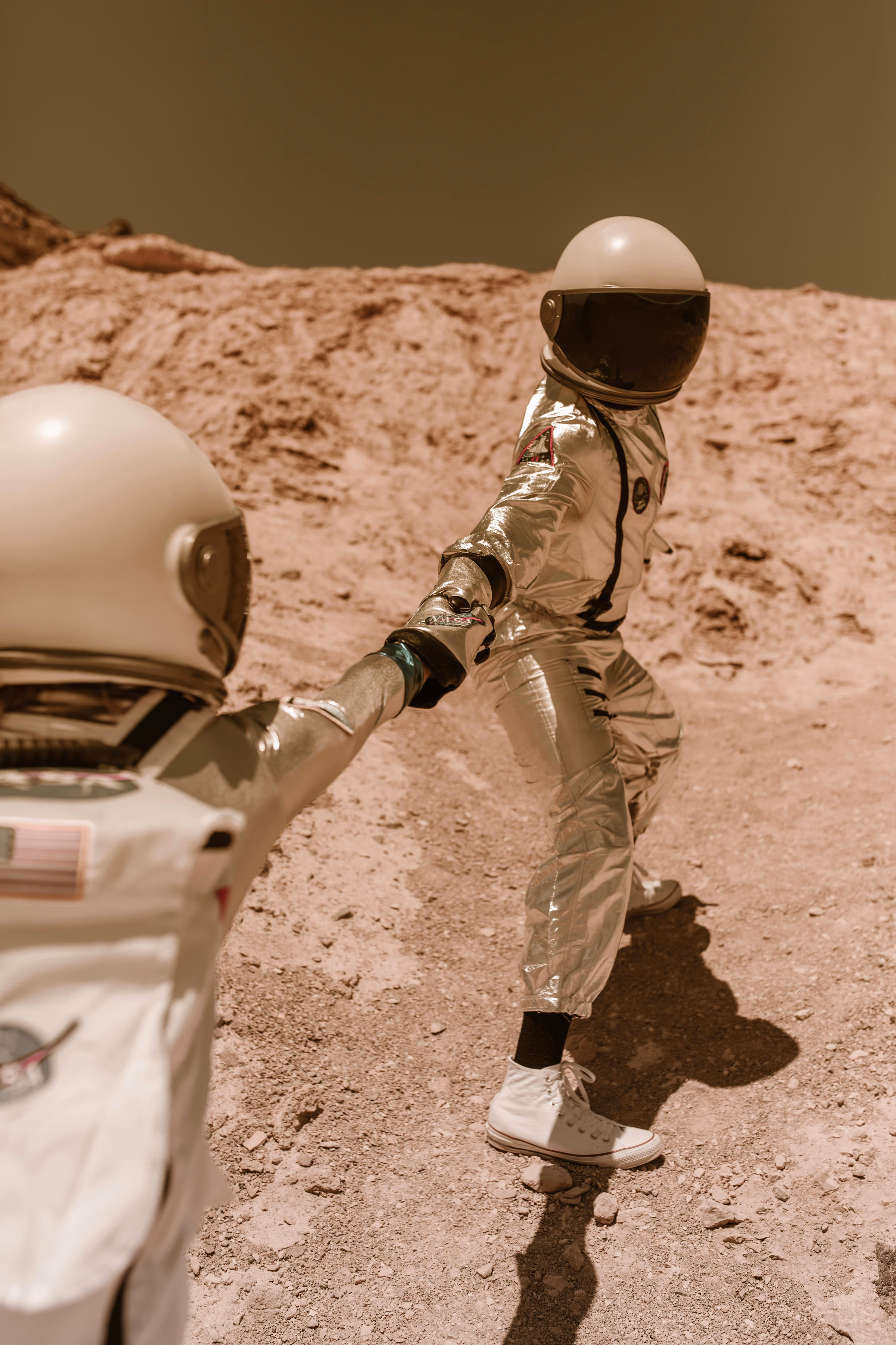 Two astronauts in reflective space suits, holding hands and walking on a rocky, barren landscape with a light brown background.