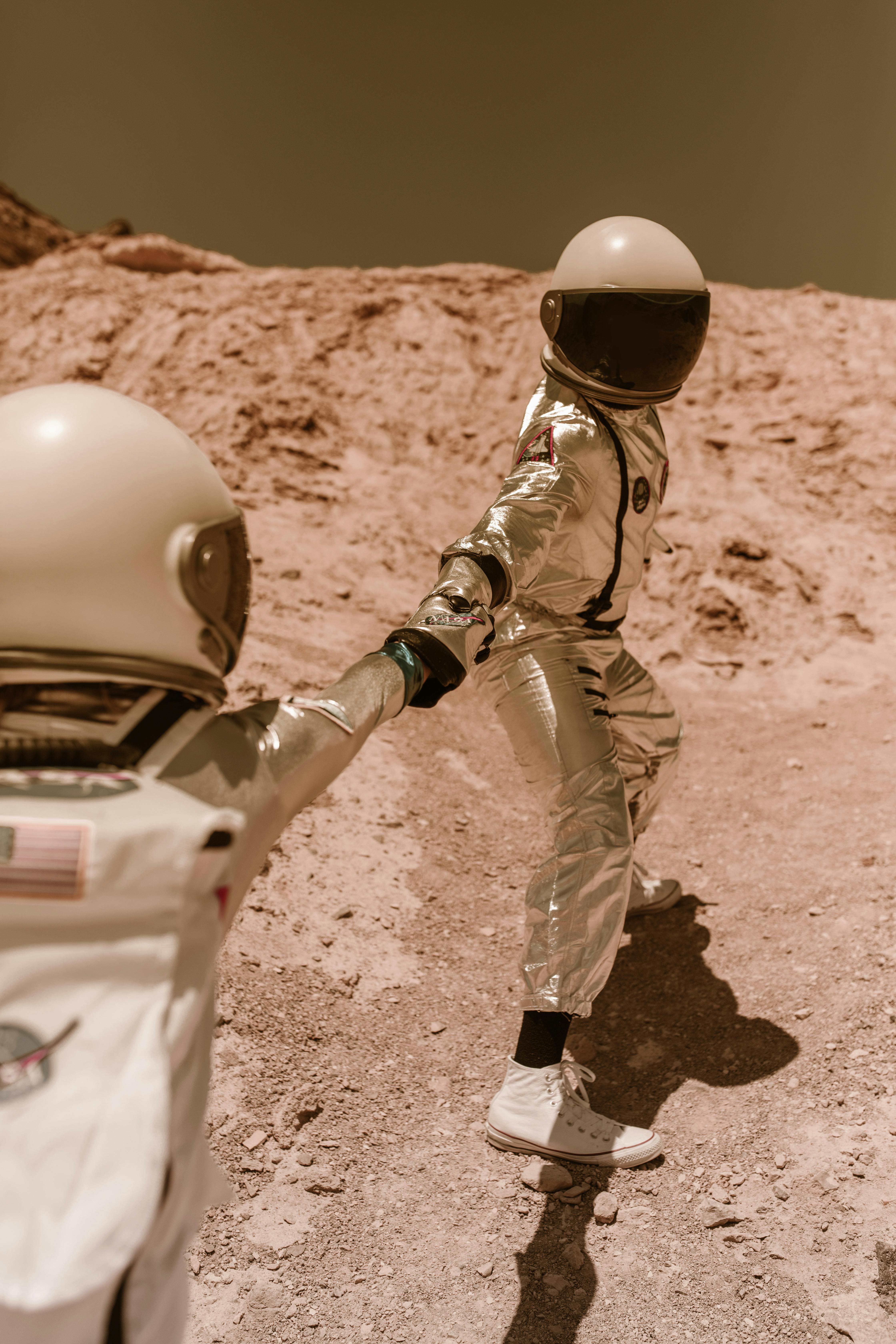 Two astronauts in reflective space suits, holding hands and walking on a rocky, barren landscape with a light brown background.