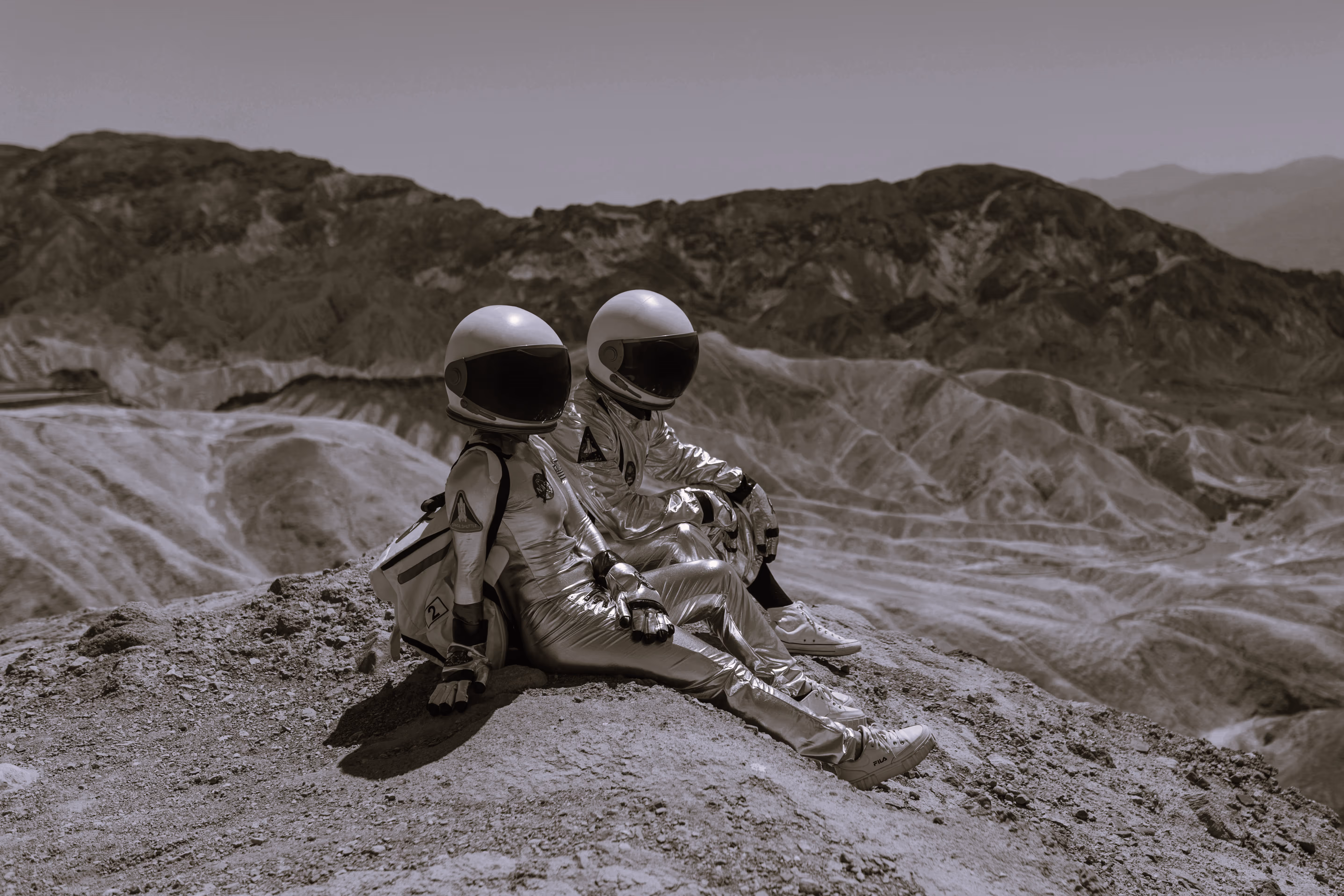 Two astronauts sitting on a rocky landscape, wearing reflective helmets and silver space suits. The background features mountainous terrain under a clear sky.