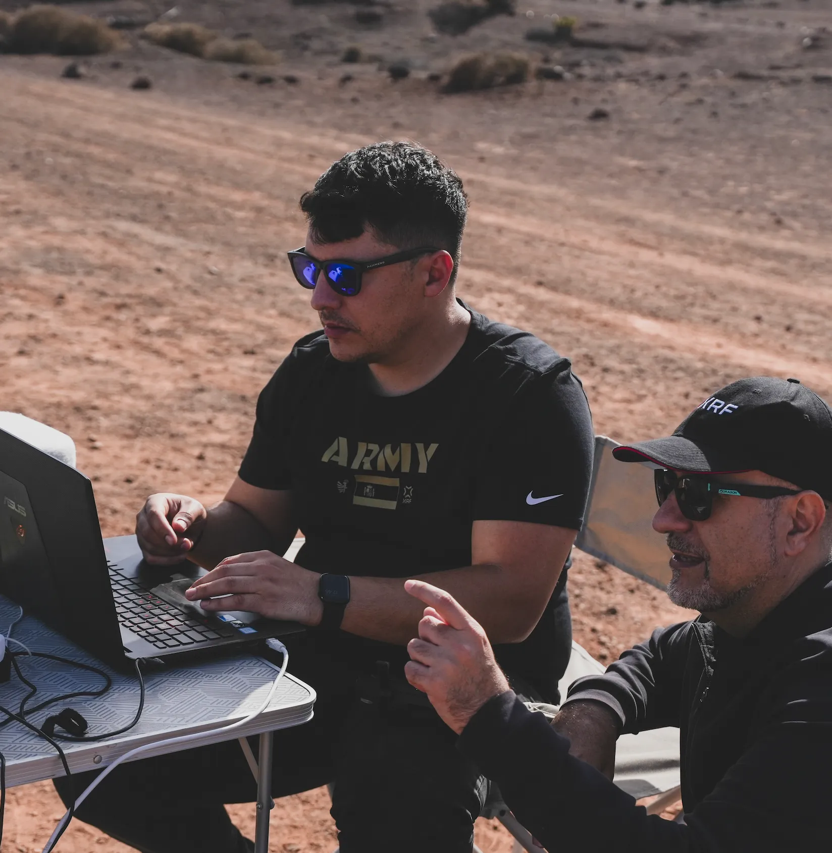 Two men working outdoors with a laptop, one pointing and the other typing