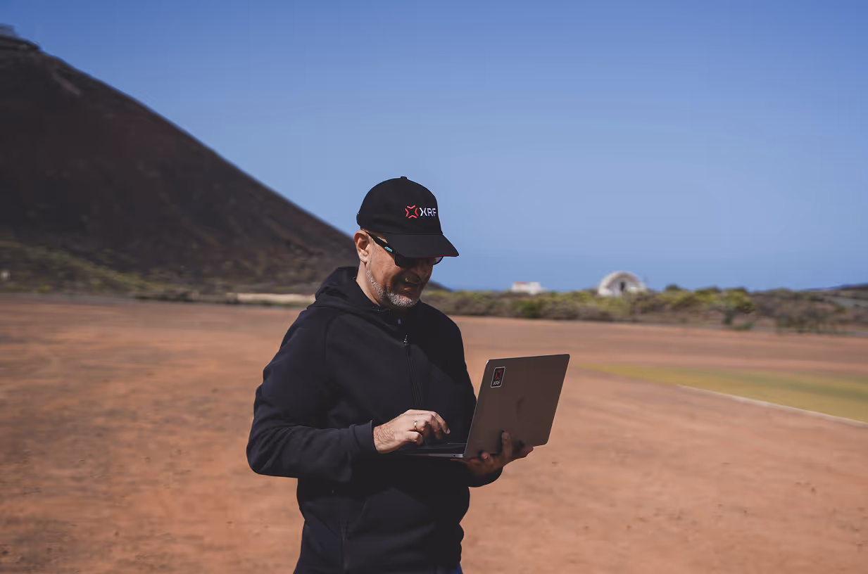 Man wearing an XRF cap and hoodie, using a laptop outdoors with a volcanic background