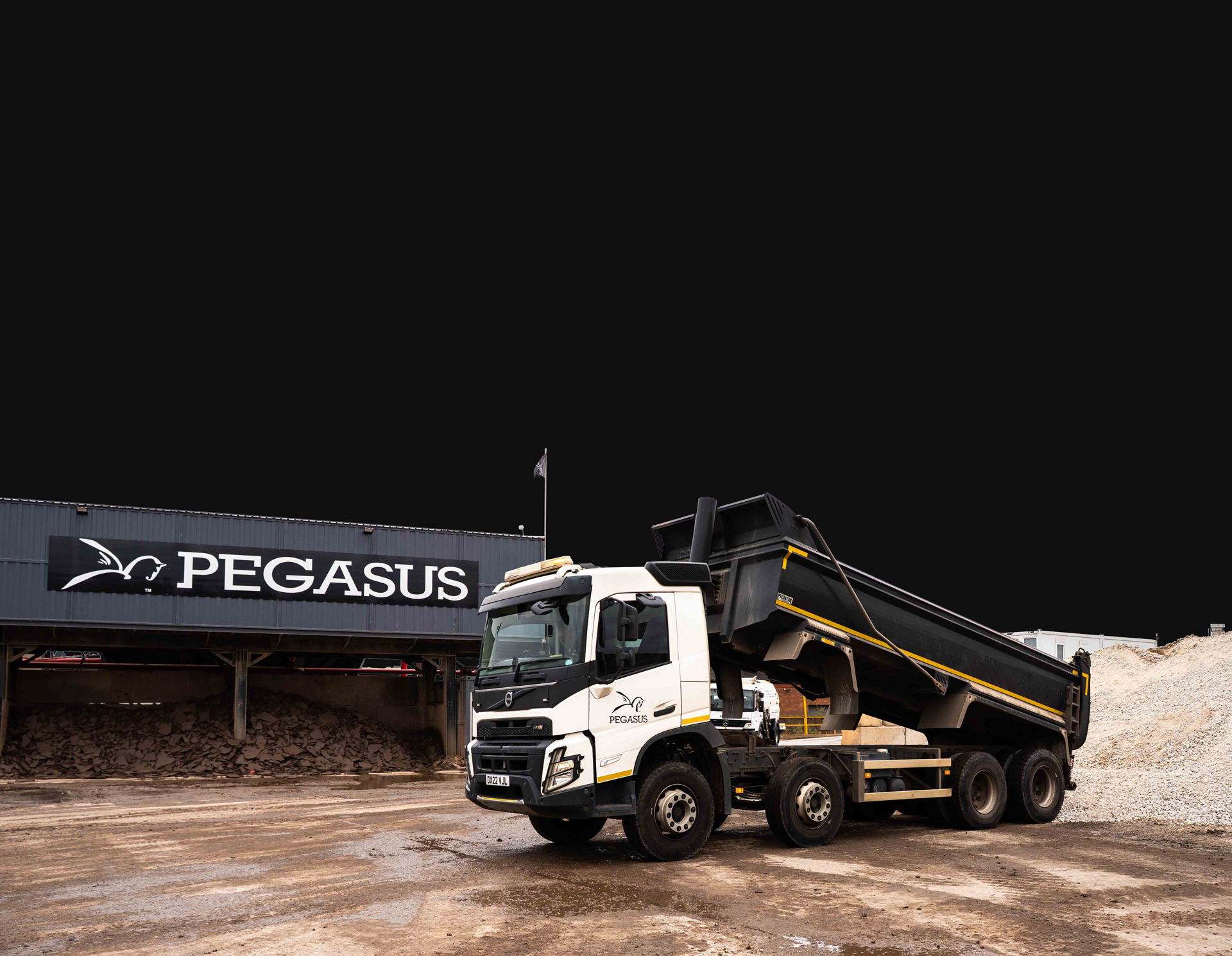 White and black Pegasus-branded dump truck with raised bed loaded with rocks at a gravel yard.
