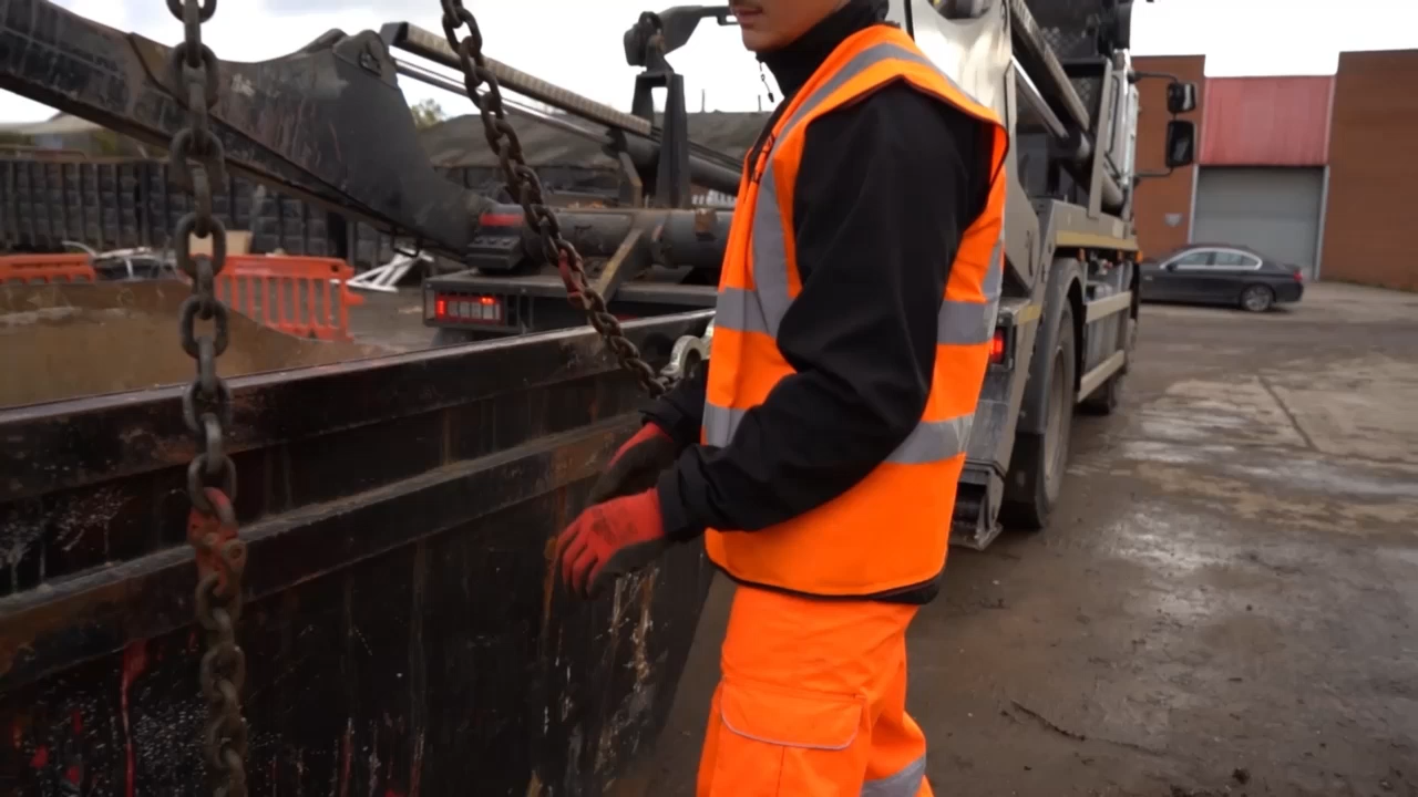 Construction worker wearing orange safety vest and gloves operating chains near heavy equipment at a muddy site.