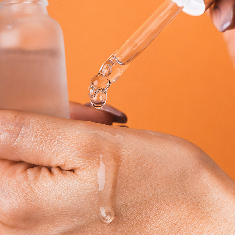 Hand hält eine Pipette, aus der ein Tropfen eines transparenten Serums auf die Haut fällt, vor orangem Hintergrund.