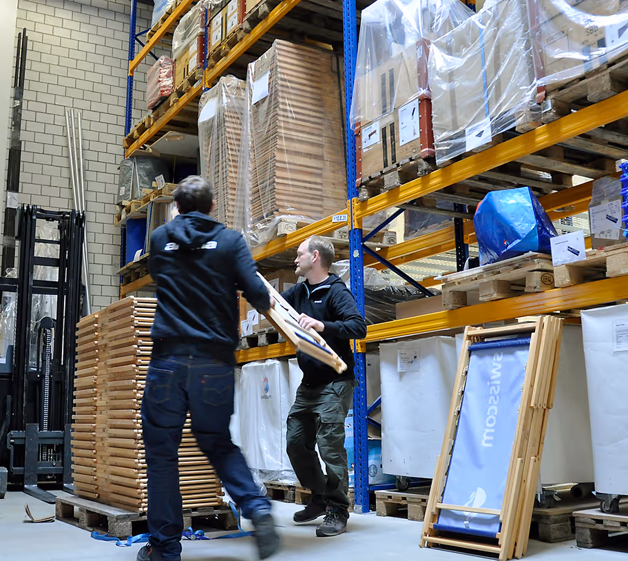 Two workers move wooden frames in a warehouse with stacked pallets and metal shelving