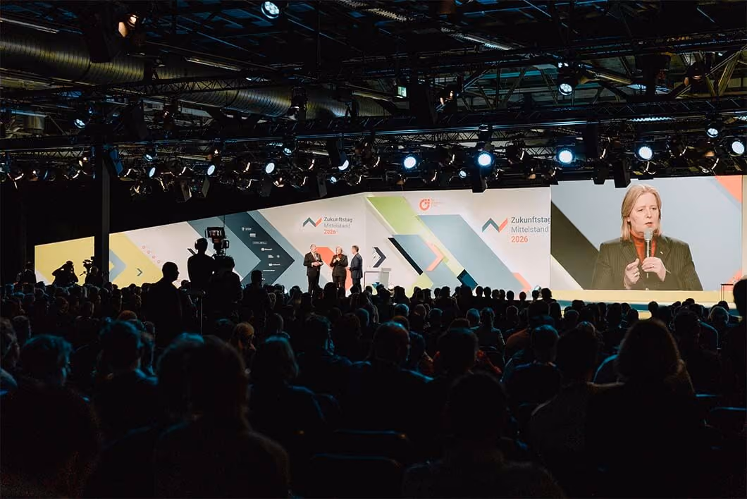 Volles Auditorium bei einer Konferenzveranstaltung mit einer großen Bühne, drei Sprechern und einer großen Leinwand, die eine Frau mit Mikrofon zeigt.