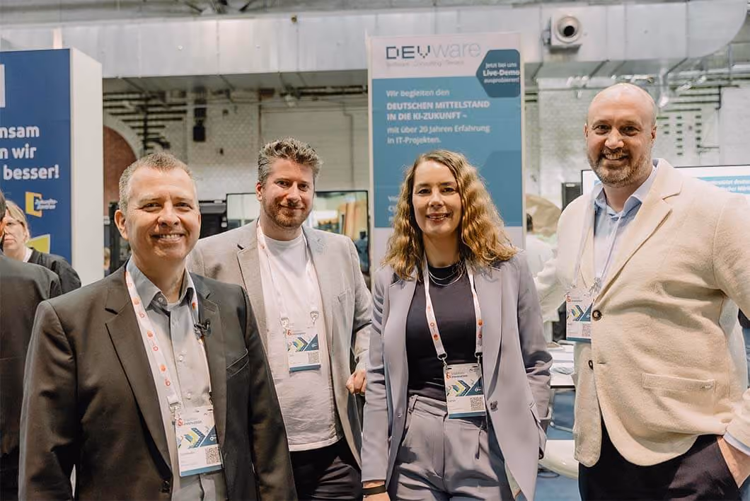Vier Personen lächeln in Business-Kleidung vor einem Messestand mit einem DEVIware-Banner im Hintergrund.