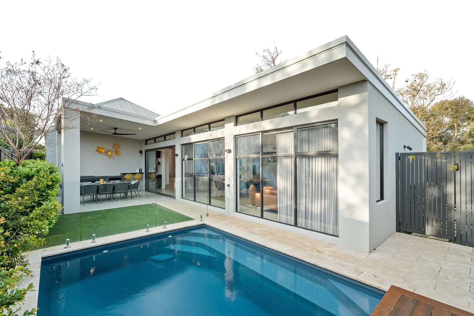 Modern house with large glass windows, an outdoor covered dining area, green lawn, and a small blue swimming pool.