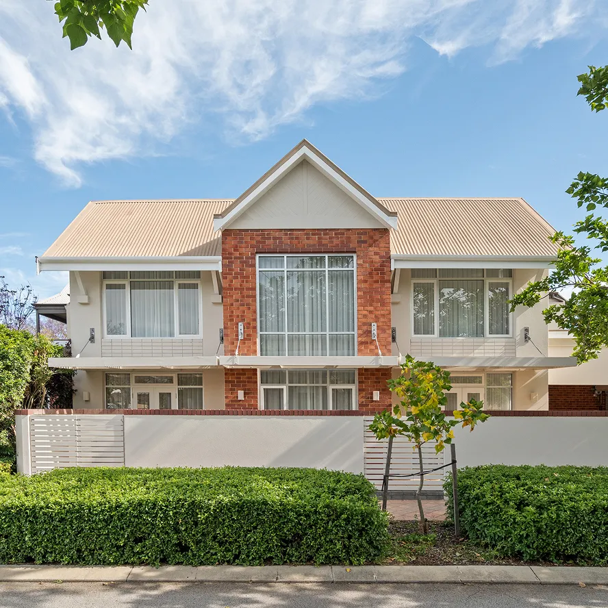 Modern two-story house with beige roof, red brick accent facade, large windows, and front garden with trimmed hedges.