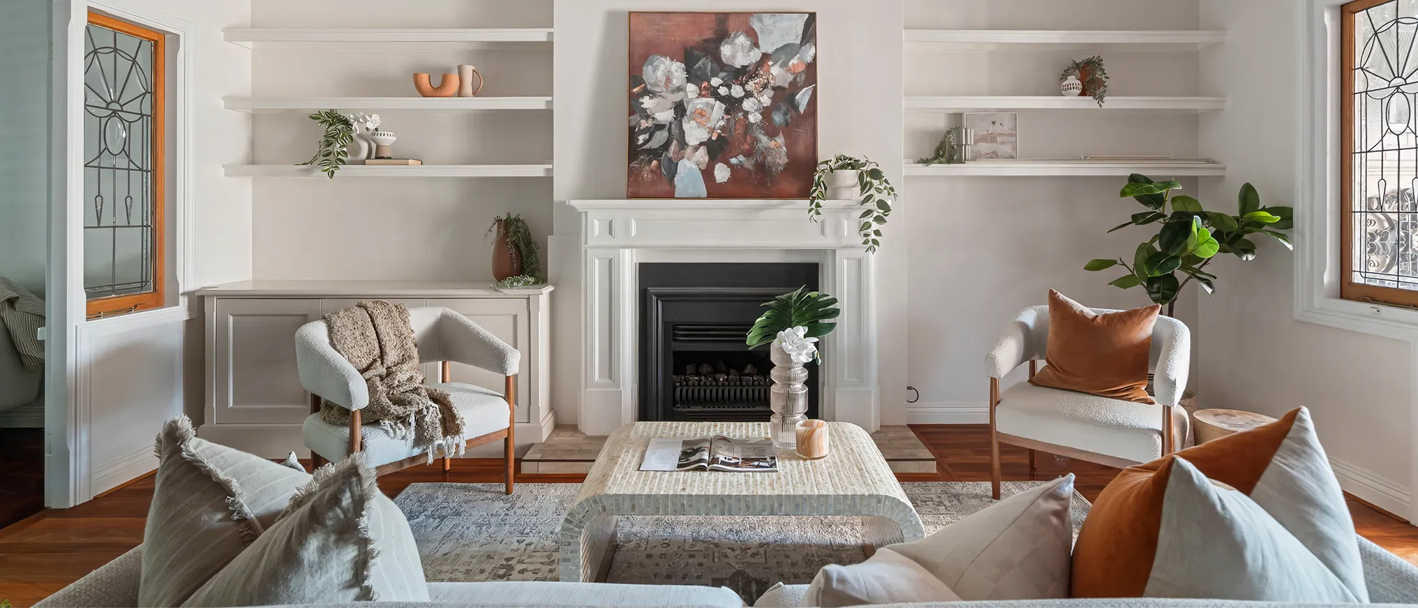 Cozy living room with white armchairs, a beige sofa with cushions, a fireplace, and decorative plants and artwork.