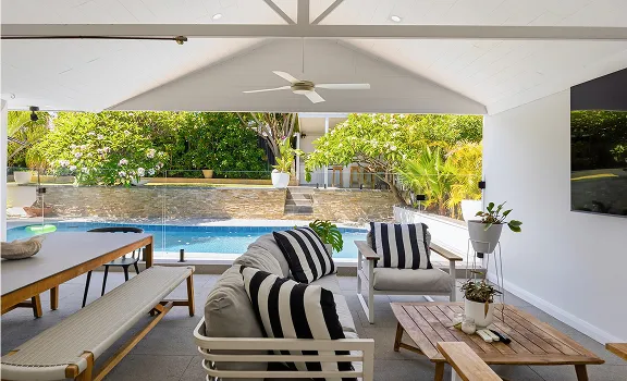 Covered outdoor patio with gray seating, striped cushions, wooden table, and view of a blue swimming pool and garden.