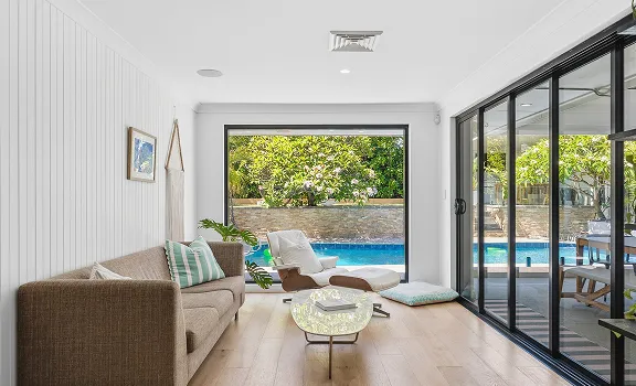 Modern living room with brown sofa, white lounge chair, glass coffee table, and large windows overlooking a backyard pool.