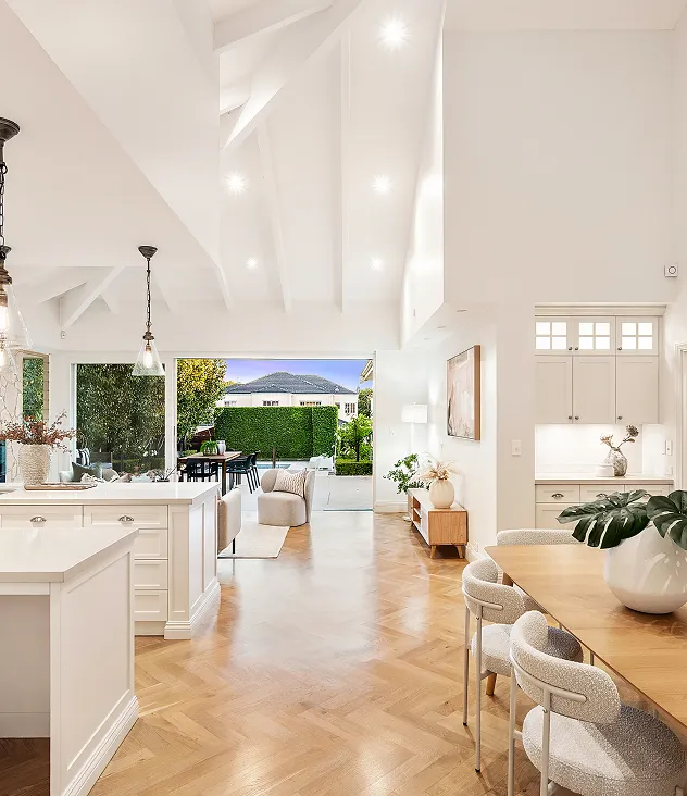 Bright open-plan living space with white kitchen cabinets, wooden dining table with modern chairs, light wood herringbone floor, and glass doors leading to an outdoor patio with greenery.