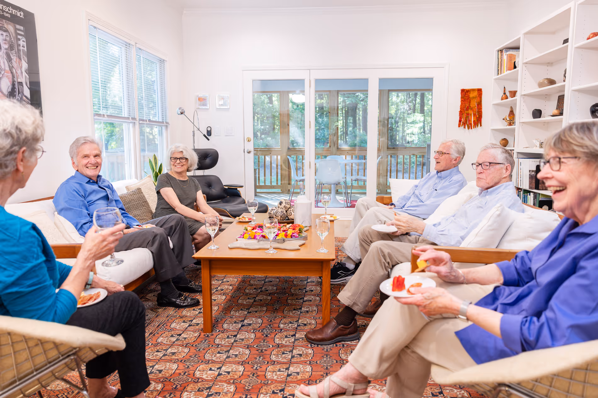 Six older adults sitting together in a living room, enjoying drinks and snacks, smiling and talking.