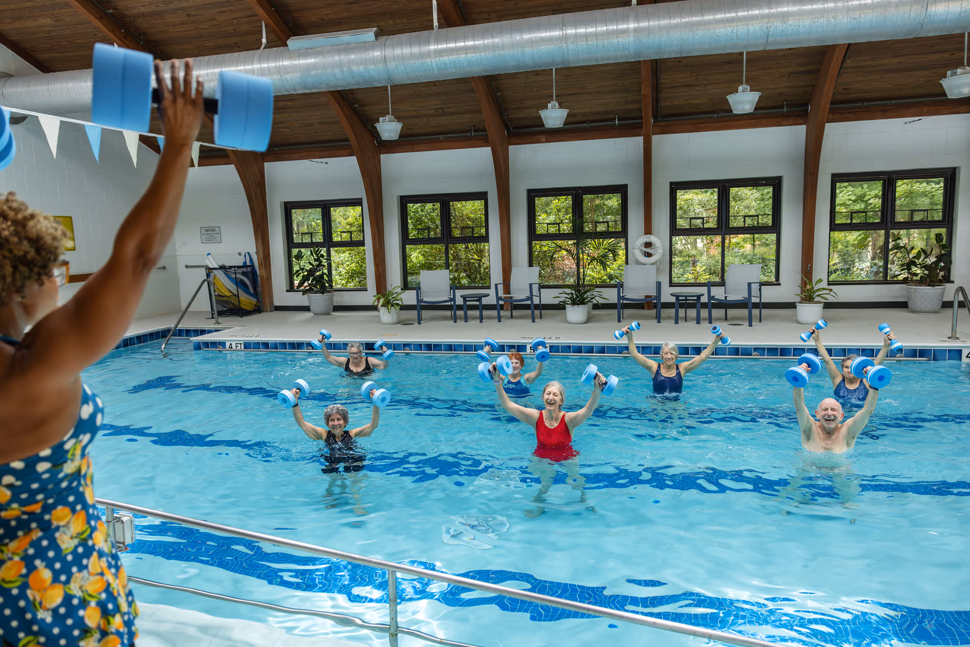 A group of people in a pool follows an instructor, raising foam dumbbells during a water exercise class.