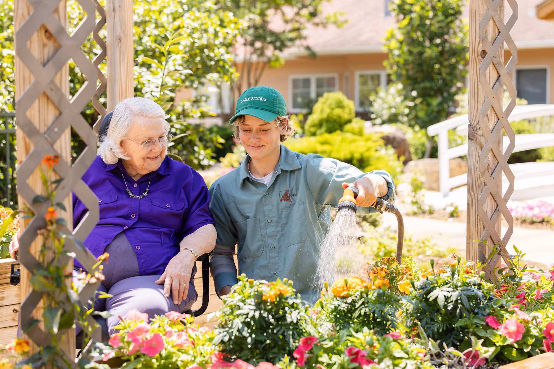 A young person in a cap waters flowers beside an older woman in a wheelchair.