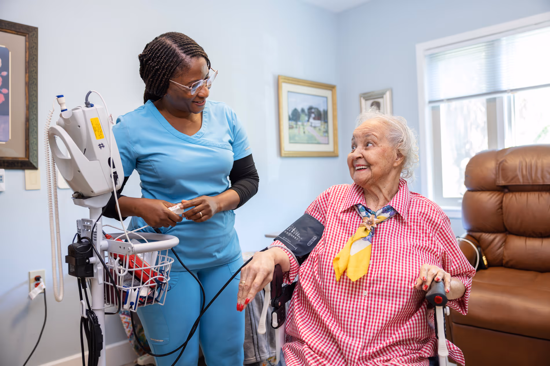 A nurse in blue scrubs measures an elderly woman's blood pressure in a cozy room.
