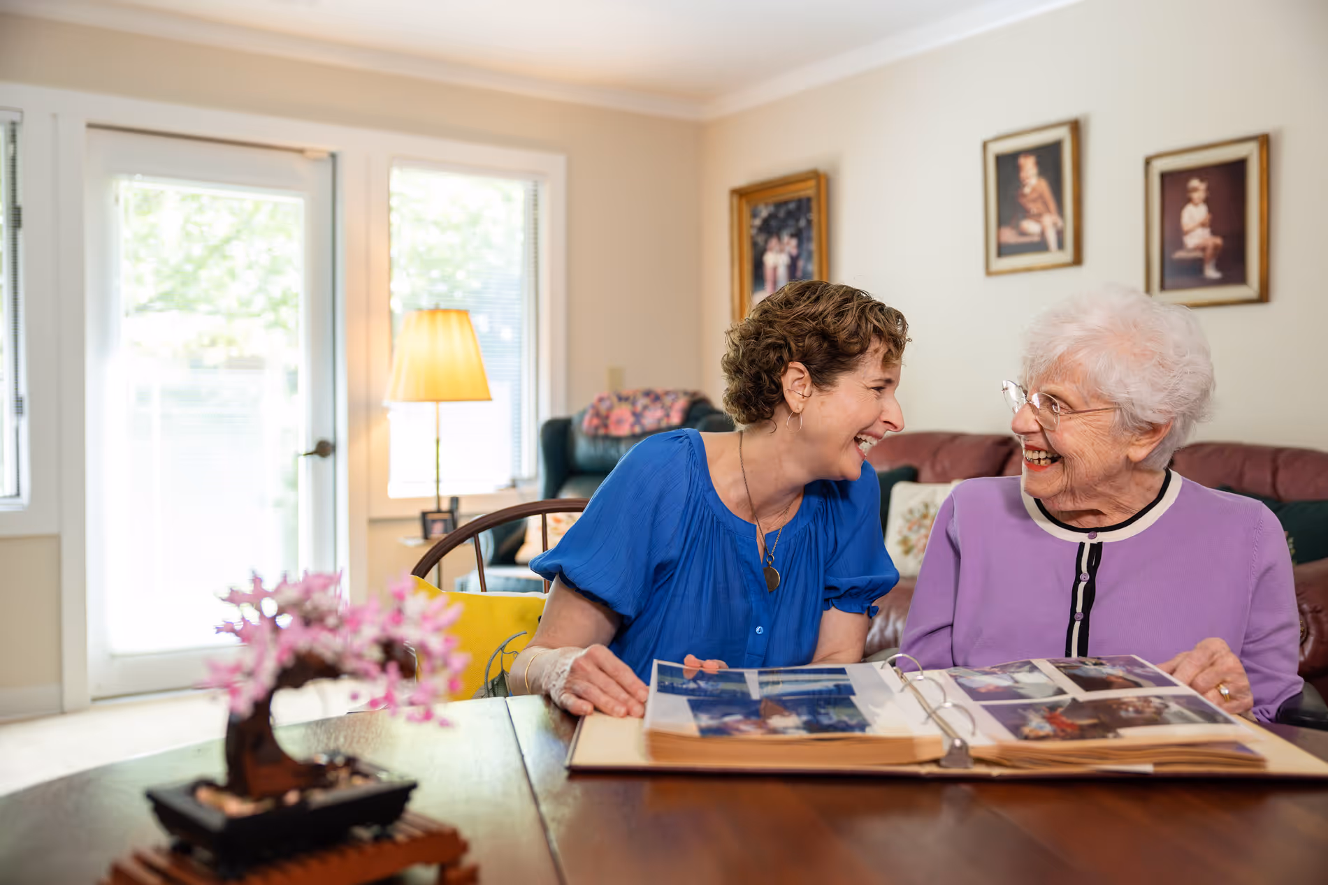 Two women sit at a table, smiling and looking through a photo album together.