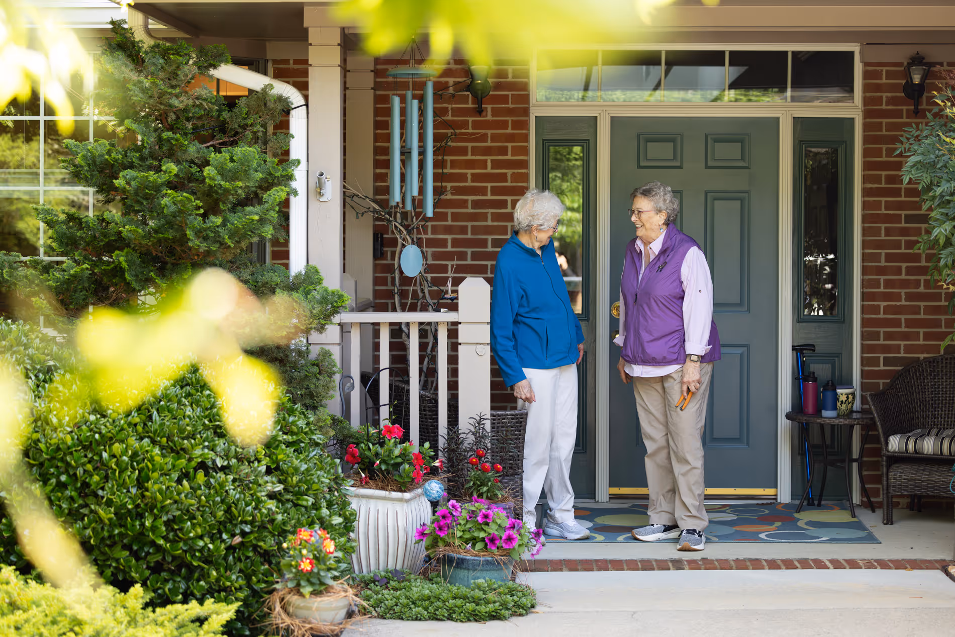 Two older adults stand talking near a front porch with flowers and a doormat.