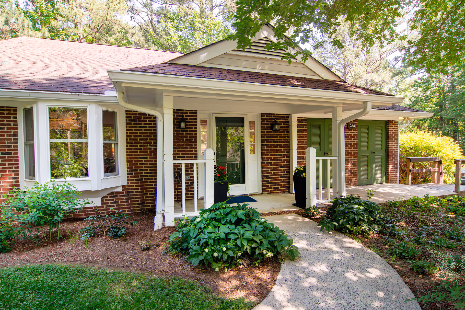 A brick house with a shaded front porch and plants along a curved walkway.