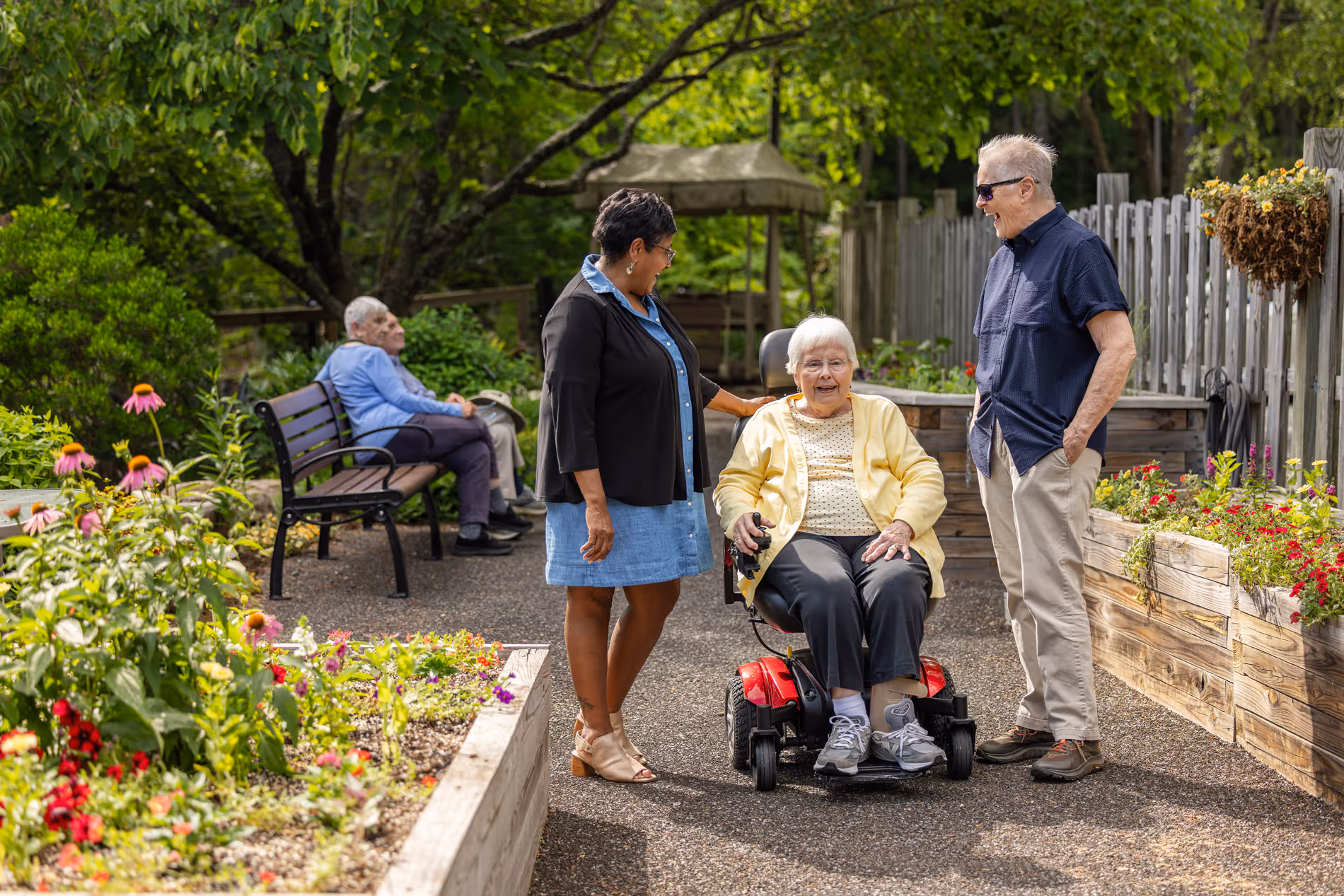 Two people talk to a woman in a wheelchair in a garden with flowers and benches.
