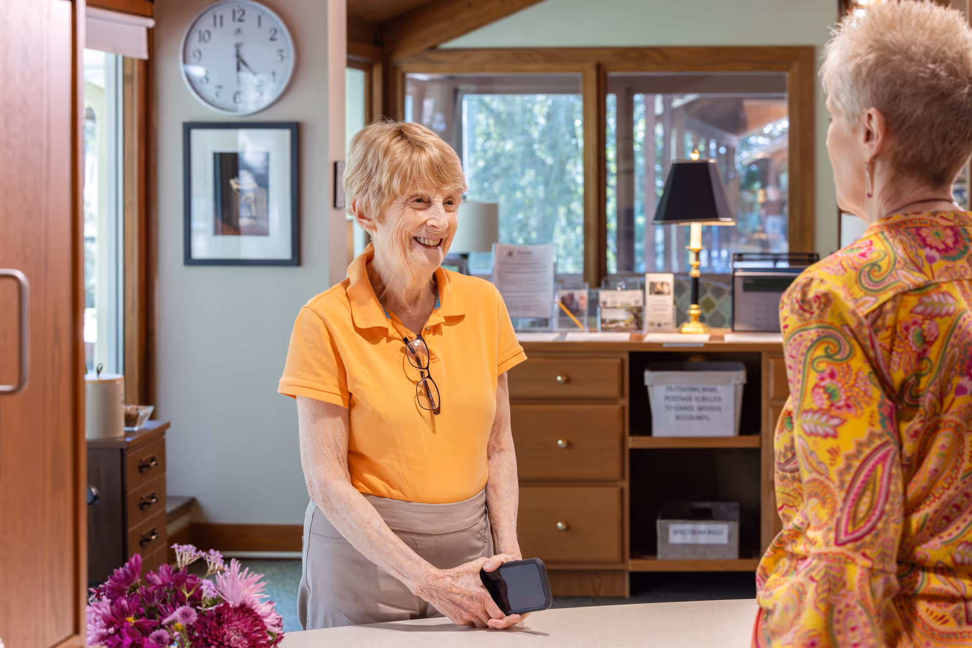 Two women talking at a reception desk, one holding a phone, with flowers nearby.
