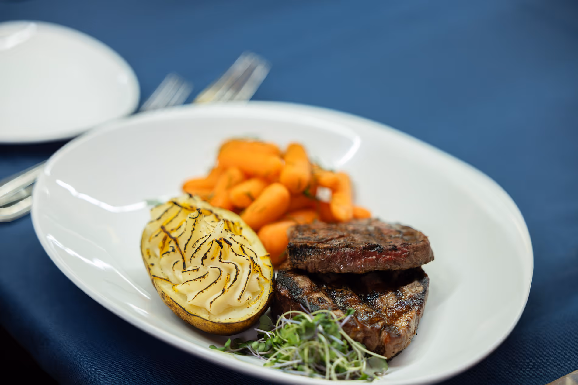 Grilled steak, roasted potatoes, and carrots on a white plate with a blue tablecloth background.