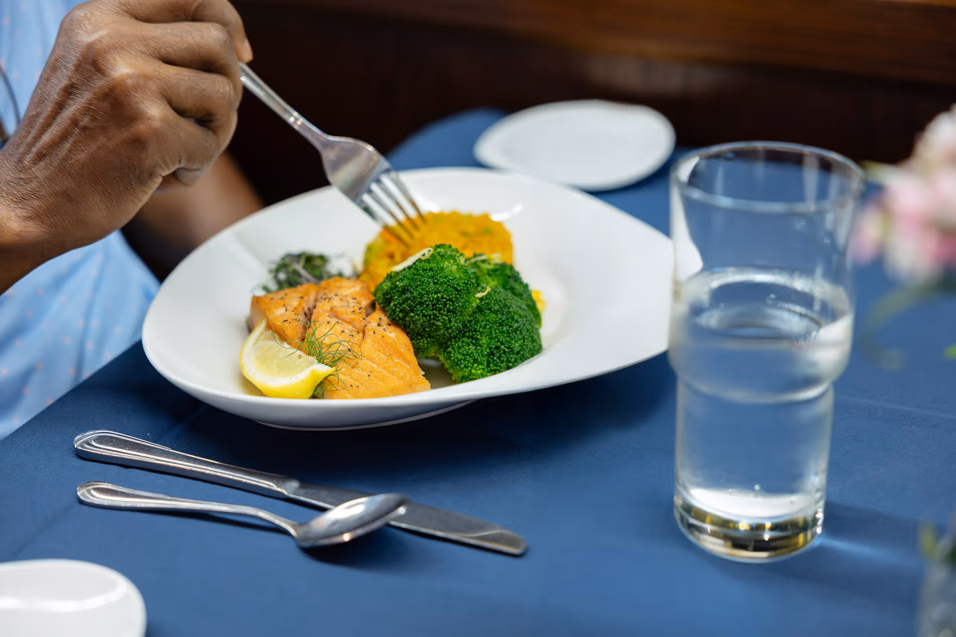 Person eating grilled salmon, broccoli, and rice at a table with a glass of water.