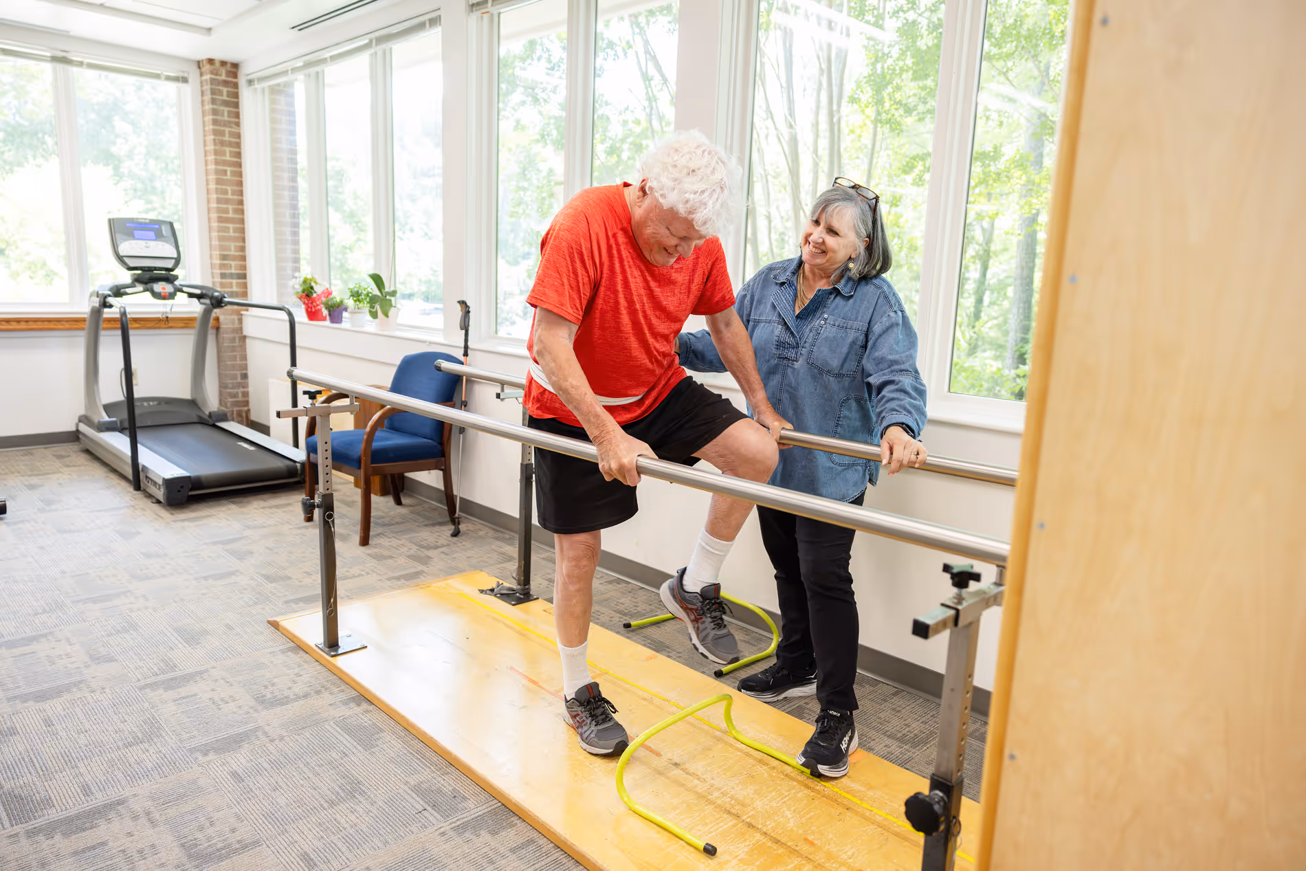 An older man practices walking on parallel bars with assistance from a woman in a therapy room.