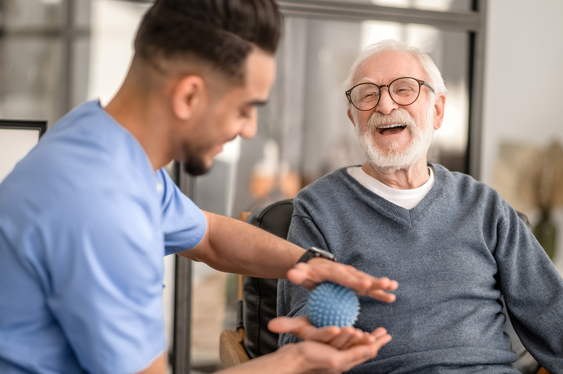 A man in a blue shirt offers an elderly man a spiky massage ball, both smiling.