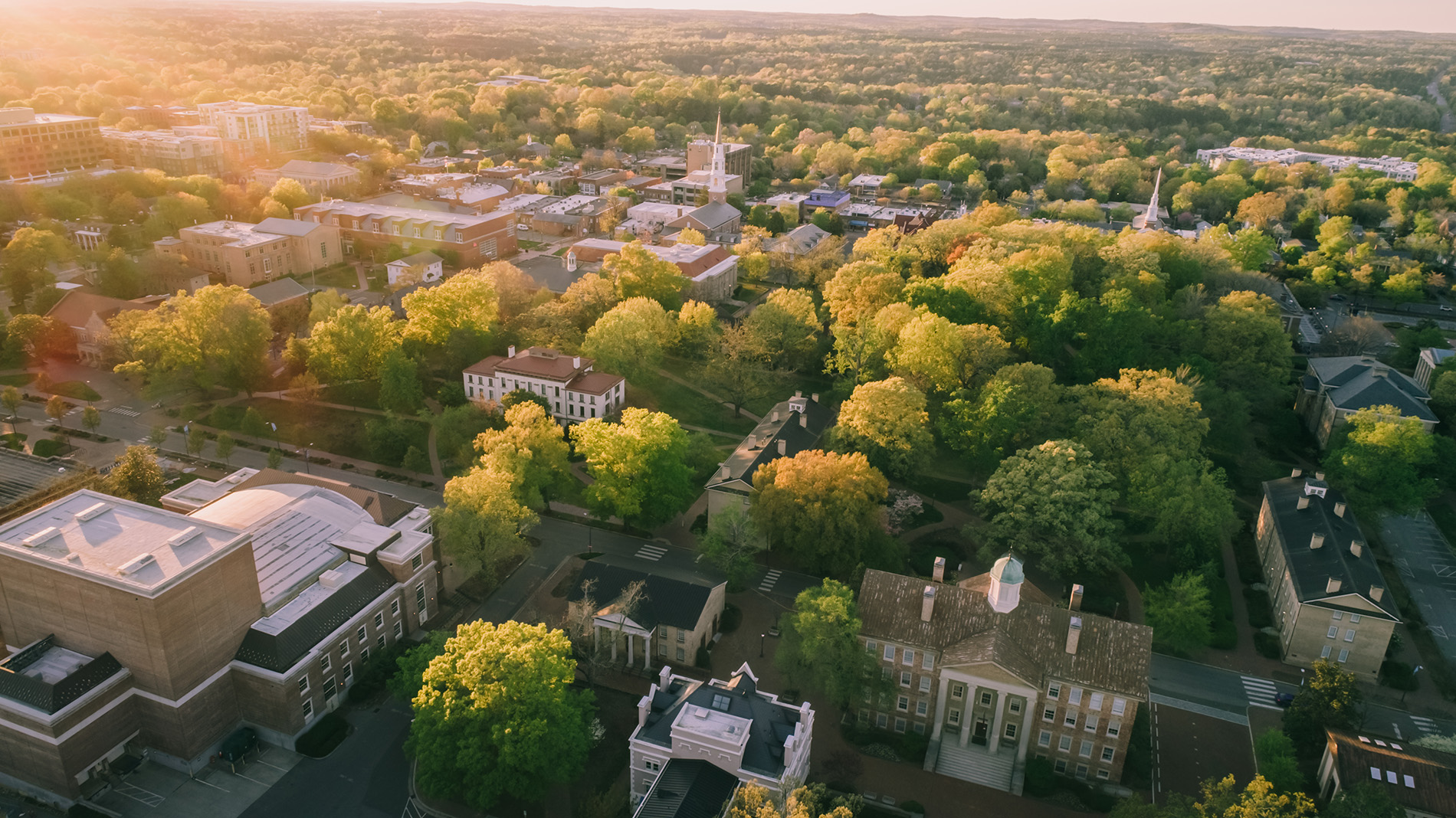 Aerial view of a campus with several buildings surrounded by many trees under a clear sky.