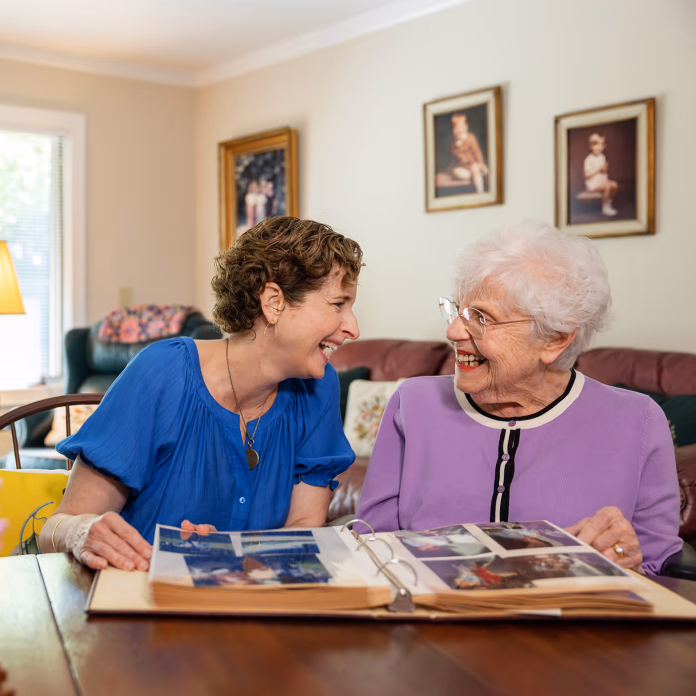 Two women smiling and looking at a photo album together in a cozy living room.