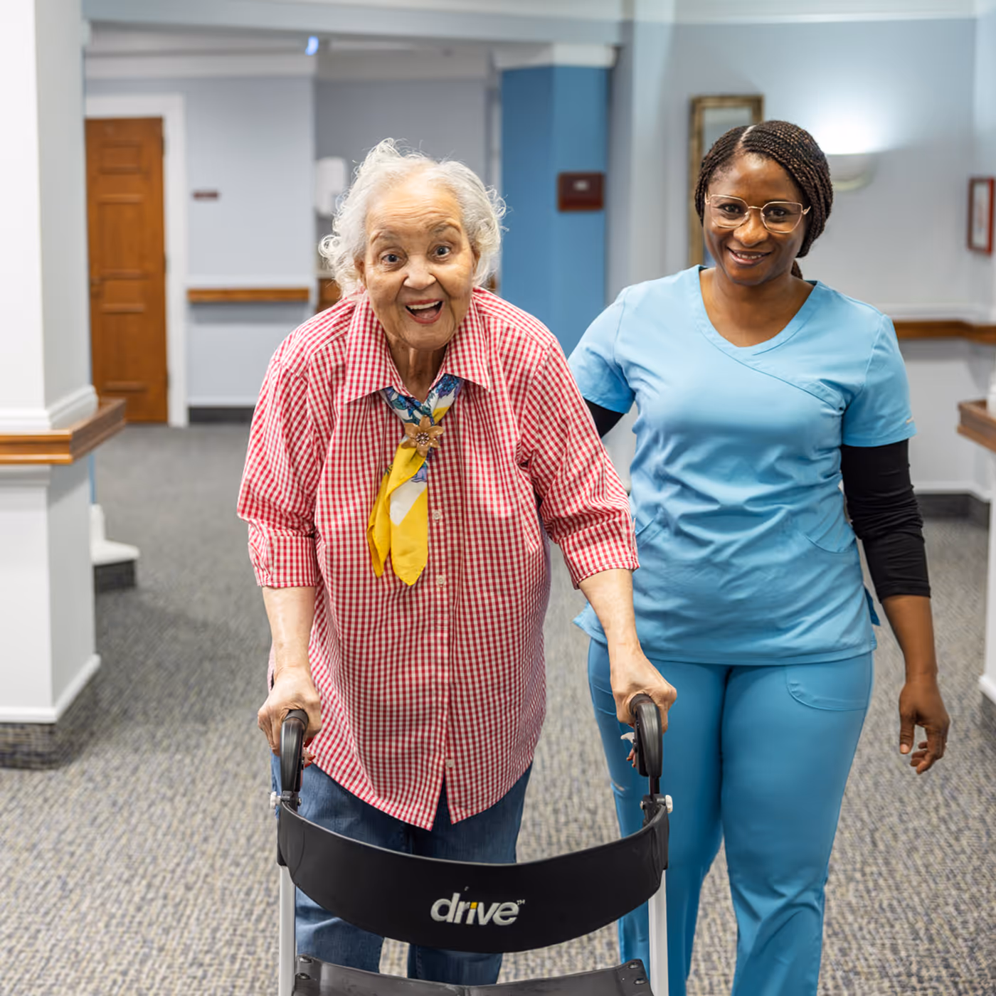 An elderly woman uses a walker, assisted by a nurse in a bright hallway.