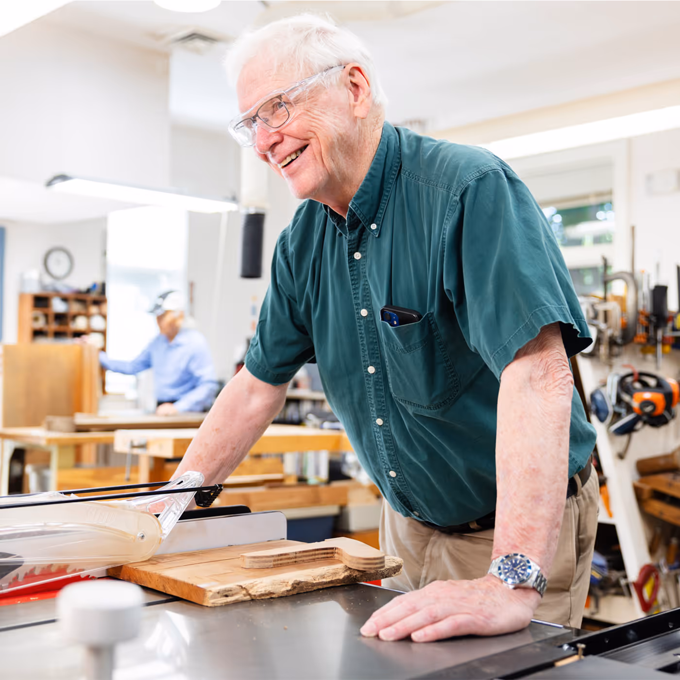Elderly man in a workshop smiling while using a table saw to cut wood.
