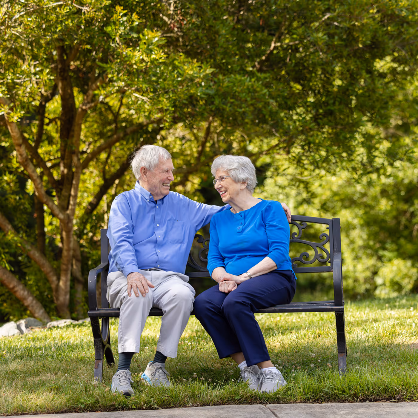 An older couple sits on a bench, smiling and chatting in a park.
