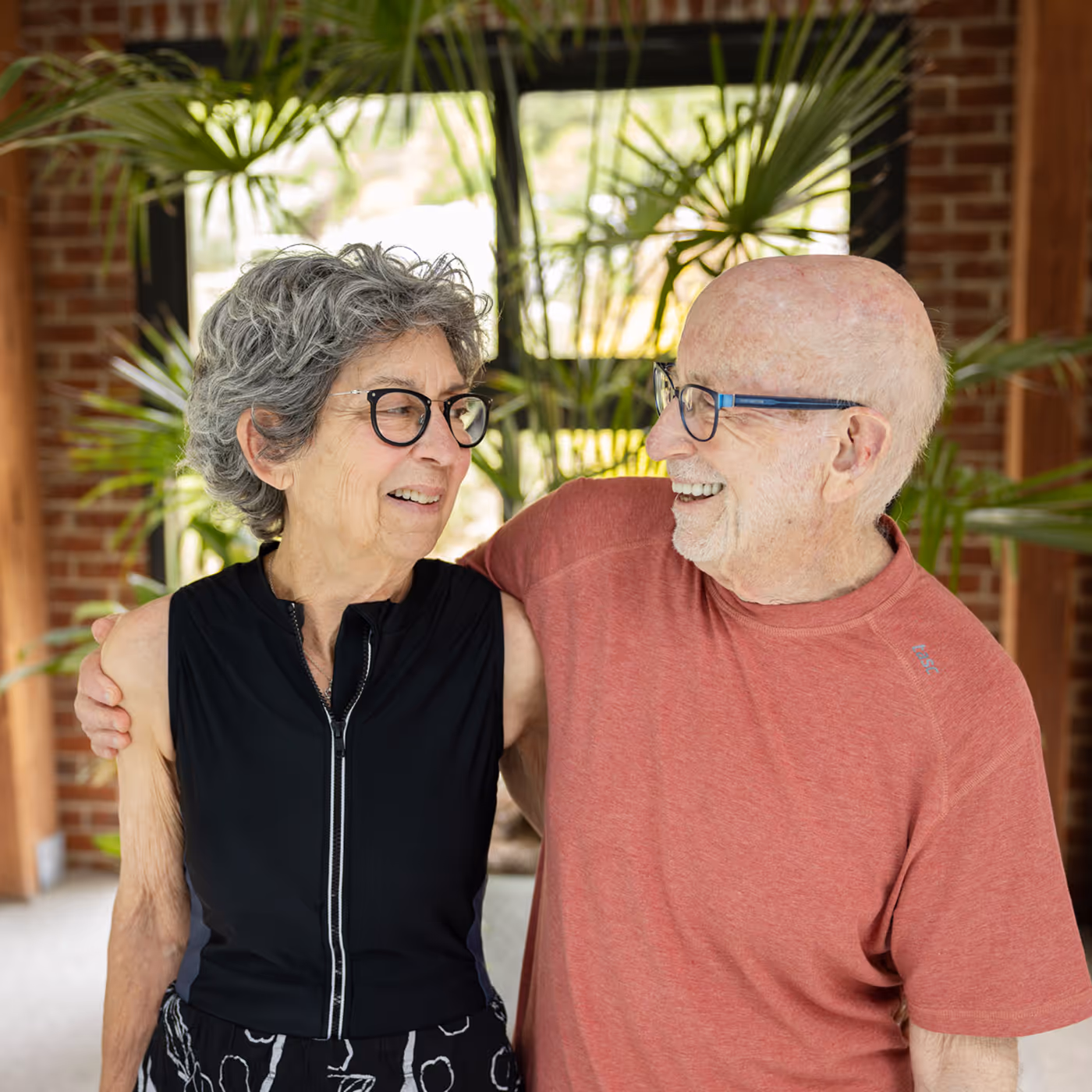 An elderly couple with glasses smiles and embraces each other warmly indoors.